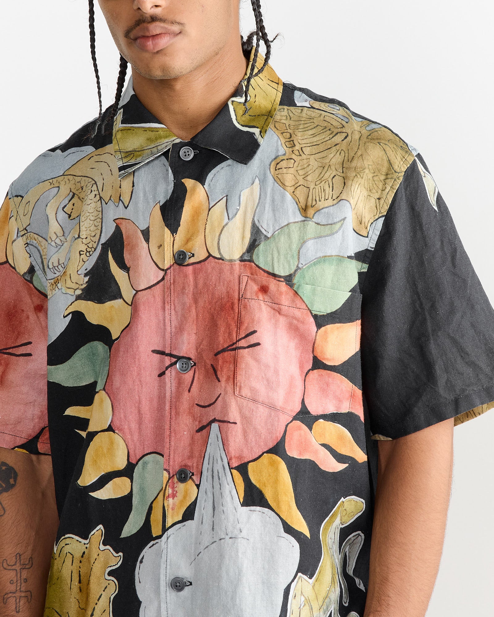 A person with braided hair wears an Our Legacy Box Shirt in Solar Power Print, showcasing a vibrant short-sleeve, boxy fit with abstract designs like a winking sun and colorful flames in red, yellow, and black hues.