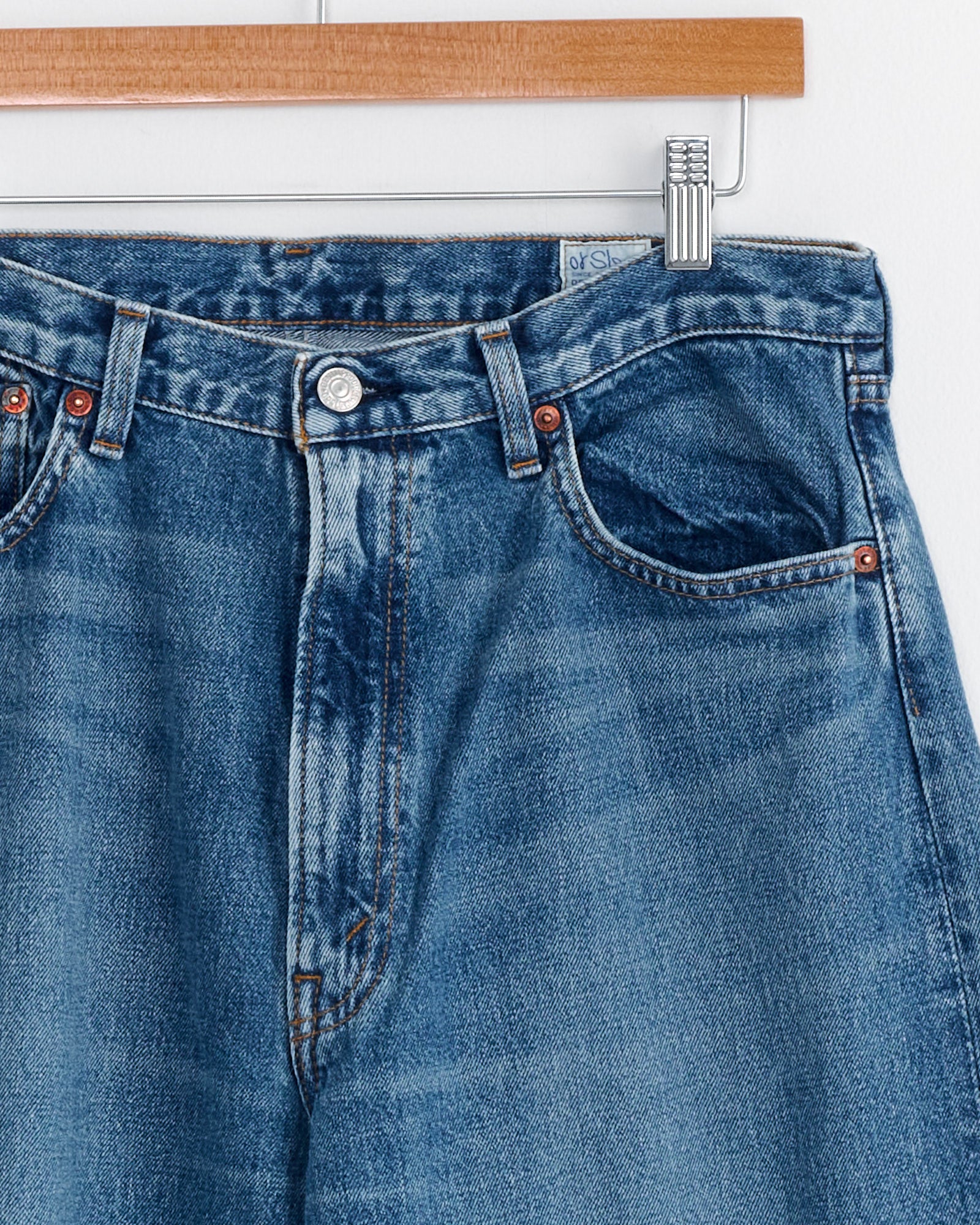 A close-up of orSlow's Super Dad's Pant in Denim Used shows the blue Japanese denim jeans on a wooden hanger, focusing on the front waistband, button, belt loops, and pocket tops against a white background.