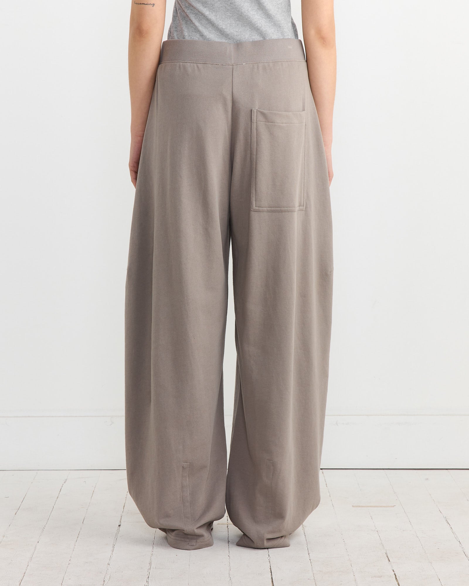 A person is seen from behind wearing Tibis Sweatshirting Winslow Pant in Dark Stone with a single back pocket. The pants complement their gray top, revealing part of an arm tattoo against a plain white wall while standing on a white wooden floor.