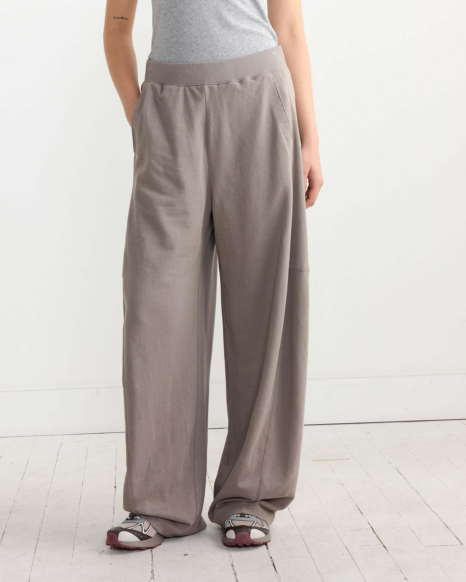 A person stands confidently in a gray top and Tibis Sweatshirting Winslow Pant in Dark Stone, hands in pockets on a white wooden floor against a plain wall. Sandals with socks highlight the relaxed look, while lightweight fabric ensures airy comfort.