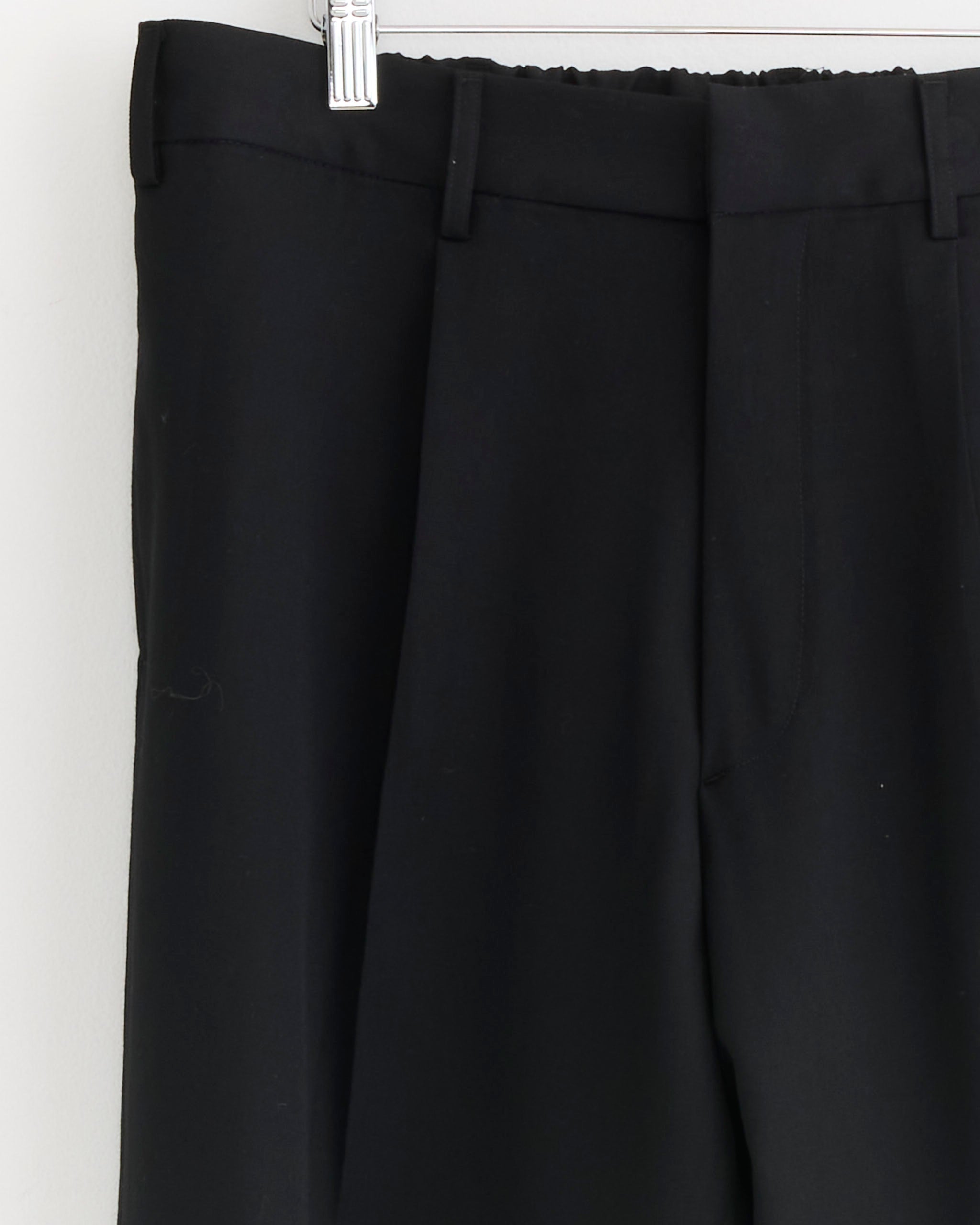 A close-up of the SMOCK West Coast Pant in Wool Gabardine Black shows the waist, belt loops, and upper section hanging on a white hanger against a light-colored wall.