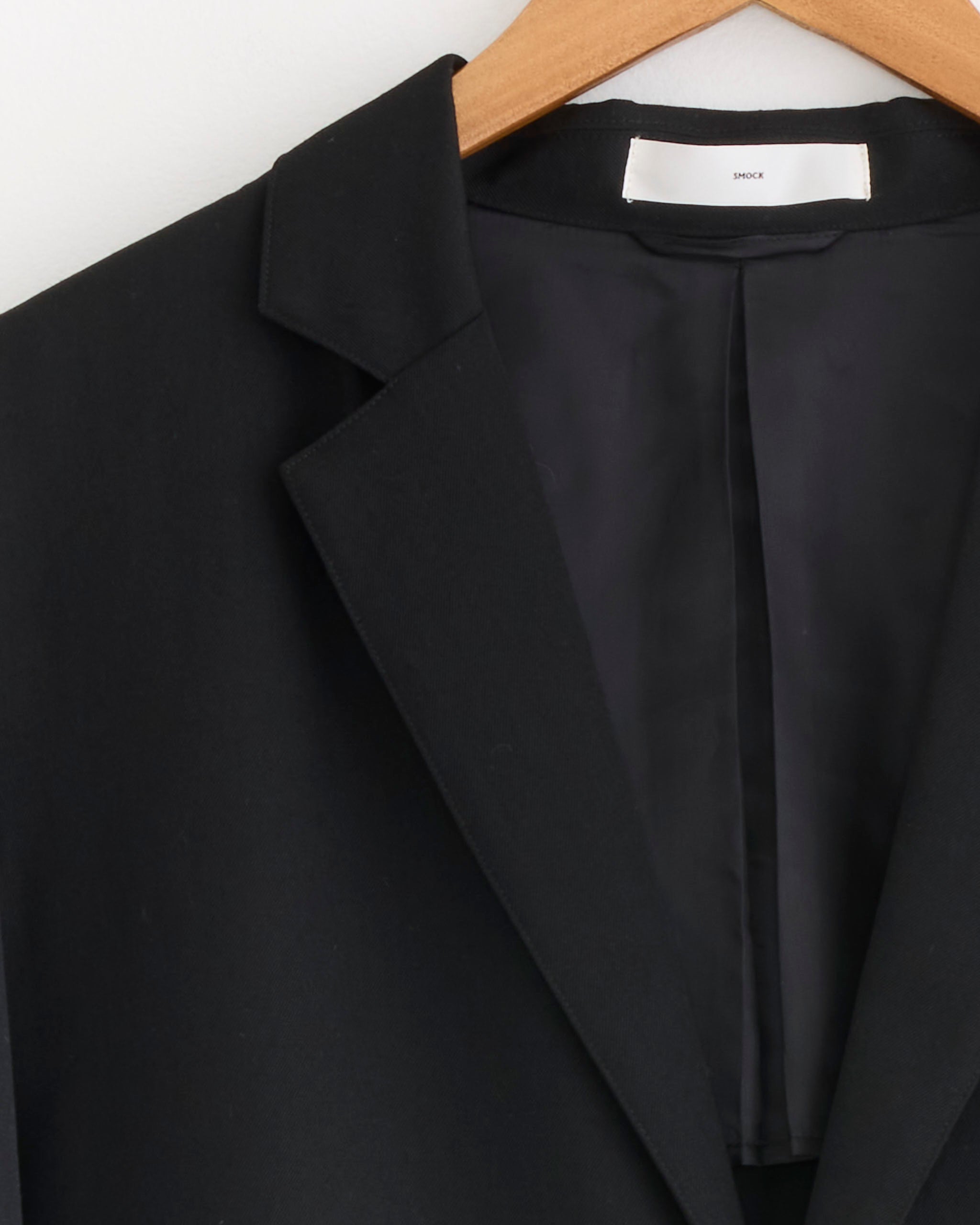 A close-up of the SMOCK Blazer in Wool Gabardine Black on a wooden hanger highlights its collar, lapel, and inner lining—an ideal choice for fall and winter.