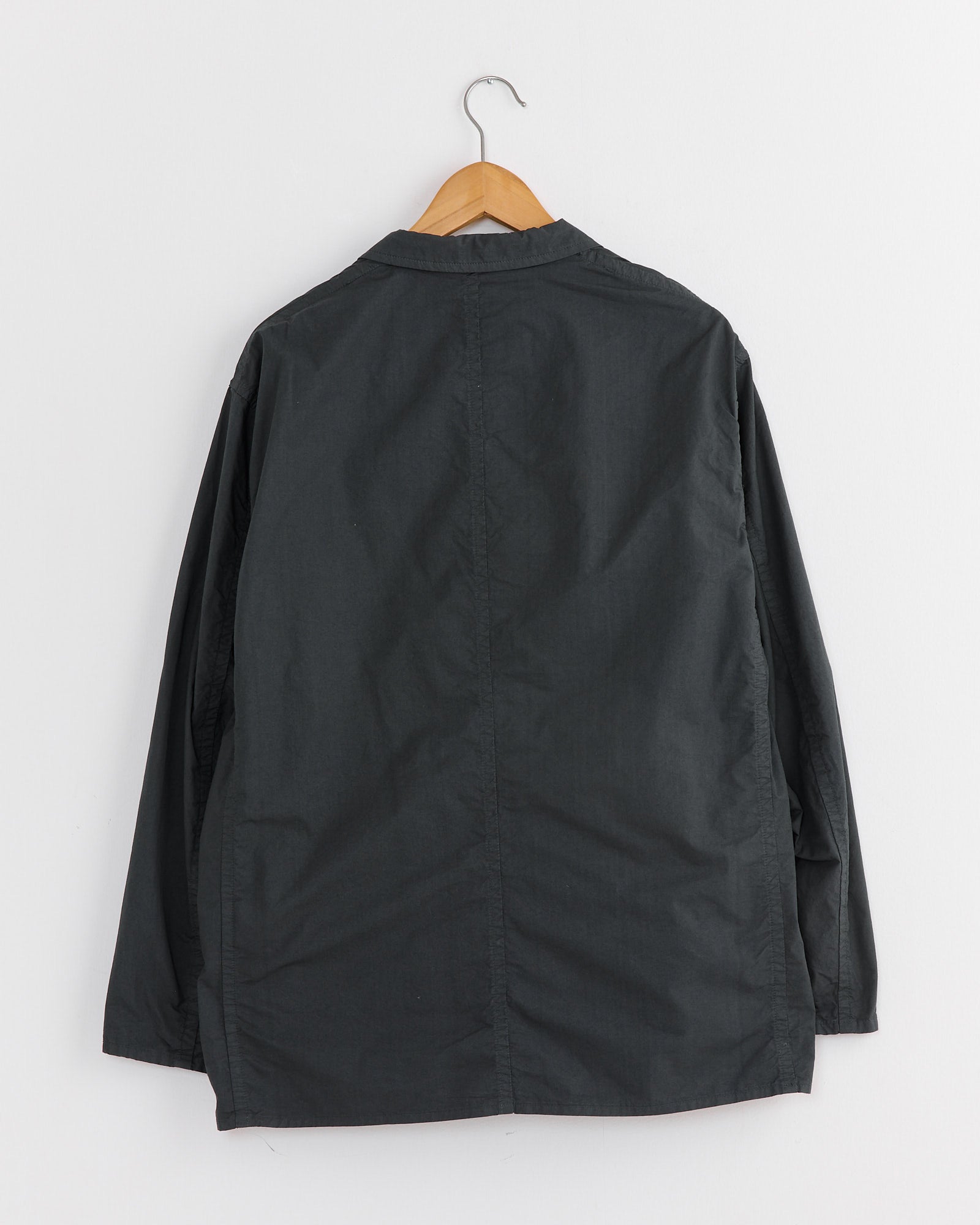 The Garment Dye 2B Jacket in Green Charcoal by Still By Hand, crafted from garment-dyed cotton, hangs on a wooden hanger against a white wall. Made in Japan with long sleeves, it is simple with no visible patterns or logos and is displayed from the back.