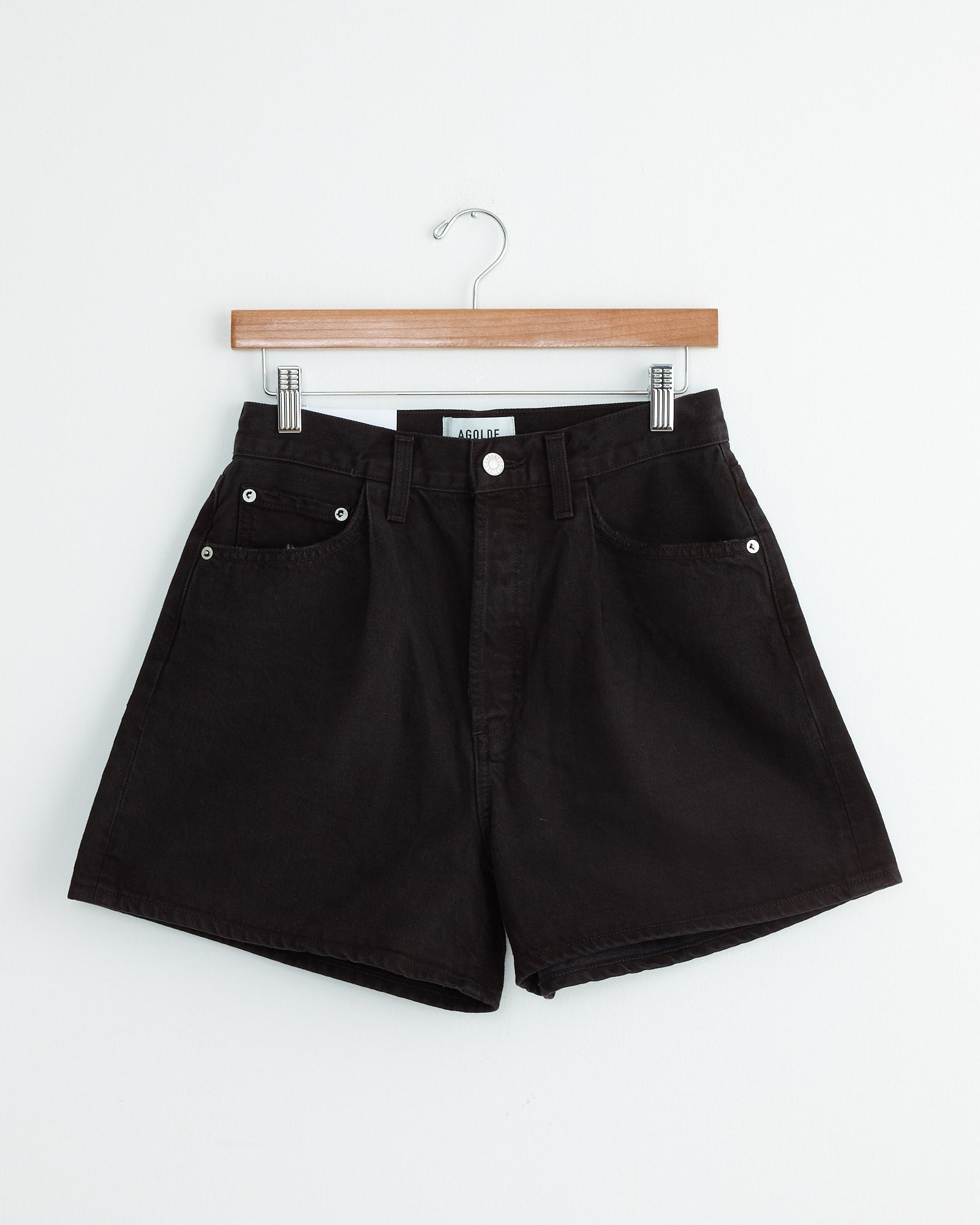 The AGOLDE Rhonda Short in Holt is a pair of high-waisted black shorts with front pockets and silver buttons, made from regenerative cotton, shown hanging on a wooden hanger against a white background.