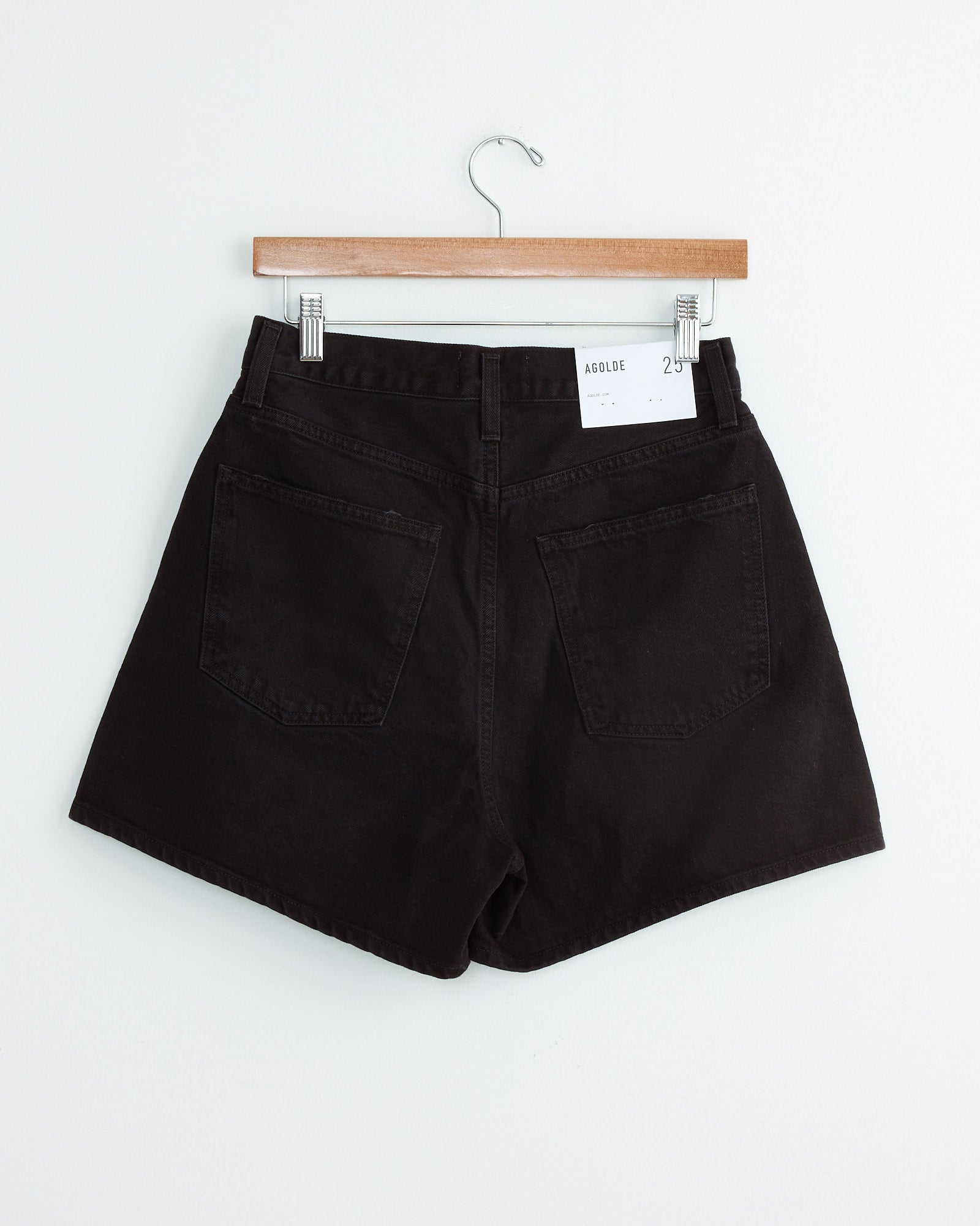 A pair of AGOLDE Rhonda Short in Holt black denim shorts hangs on a wooden hanger against a white wall, displaying the back with two pockets and a white label on the waistband.