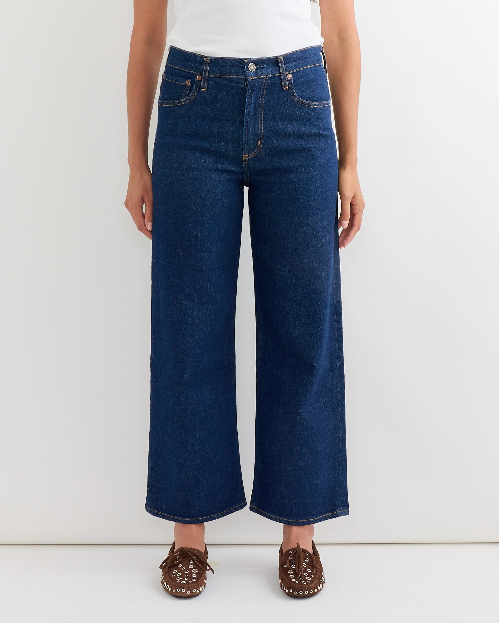 Model wearing AGOLDE Ren Jean in Reissue, a dark blue high-rise cropped sustainable denim, paired with a white top and brown woven shoes. The image highlights the lower body against a plain white background.