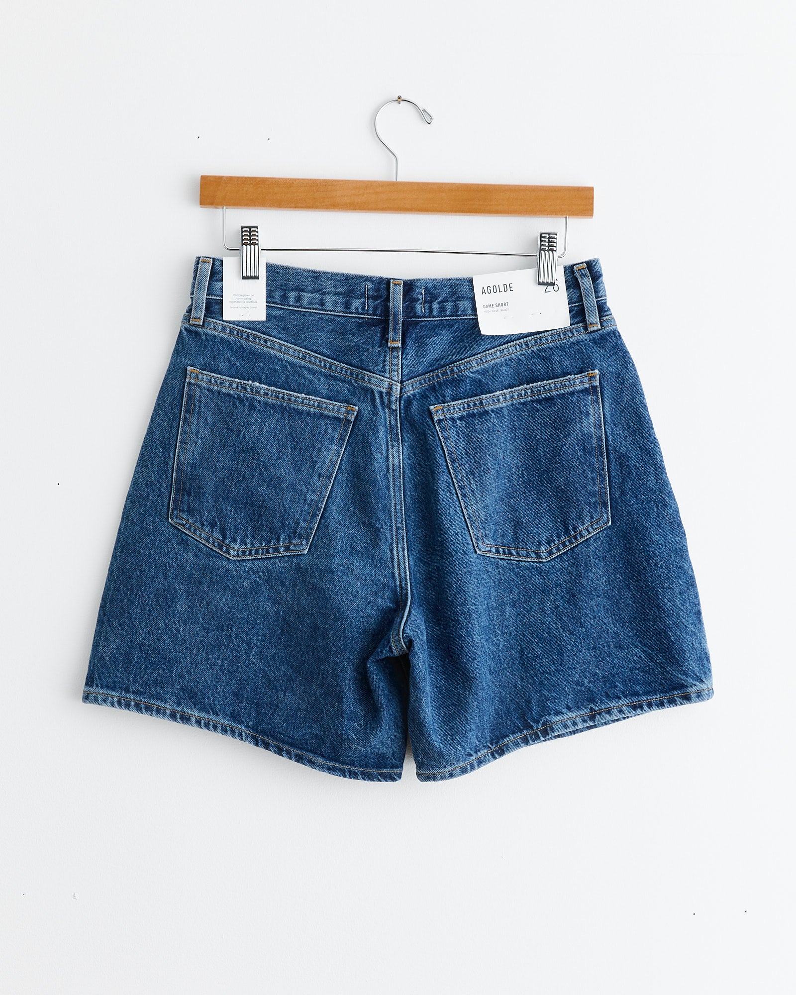 The Dame Short in Enamour by AGOLDE hangs on a wooden hanger against a white wall, displayed from the back with two large pockets and a white label on the waistband.