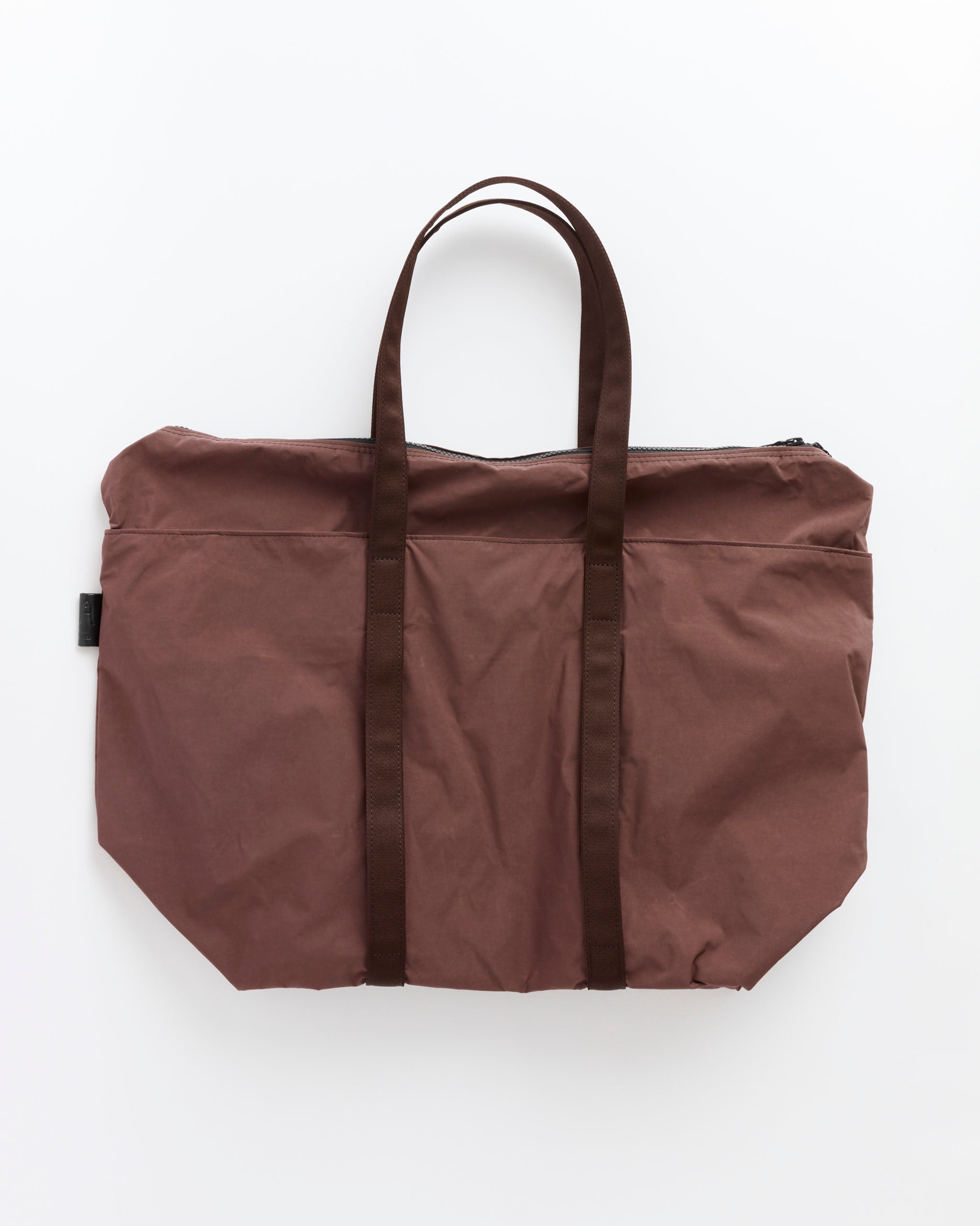 The Gabardine Tote in Bordeaux Brown by Amiacalva lies flat on a white background, featuring two dark brown handles, a top zipper closure, and a small fabric tag on one side.
