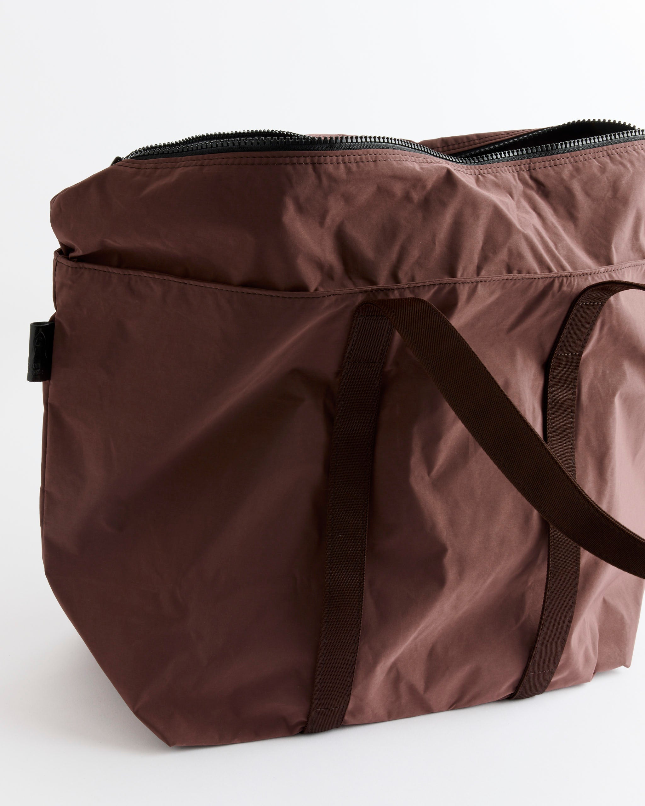 The Gabardine Tote in Bordeaux Brown by Amiacalva is a spacious tote bag crafted from textured gabardine fabric, featuring black handles and a zipper closure, shown against a plain white background.