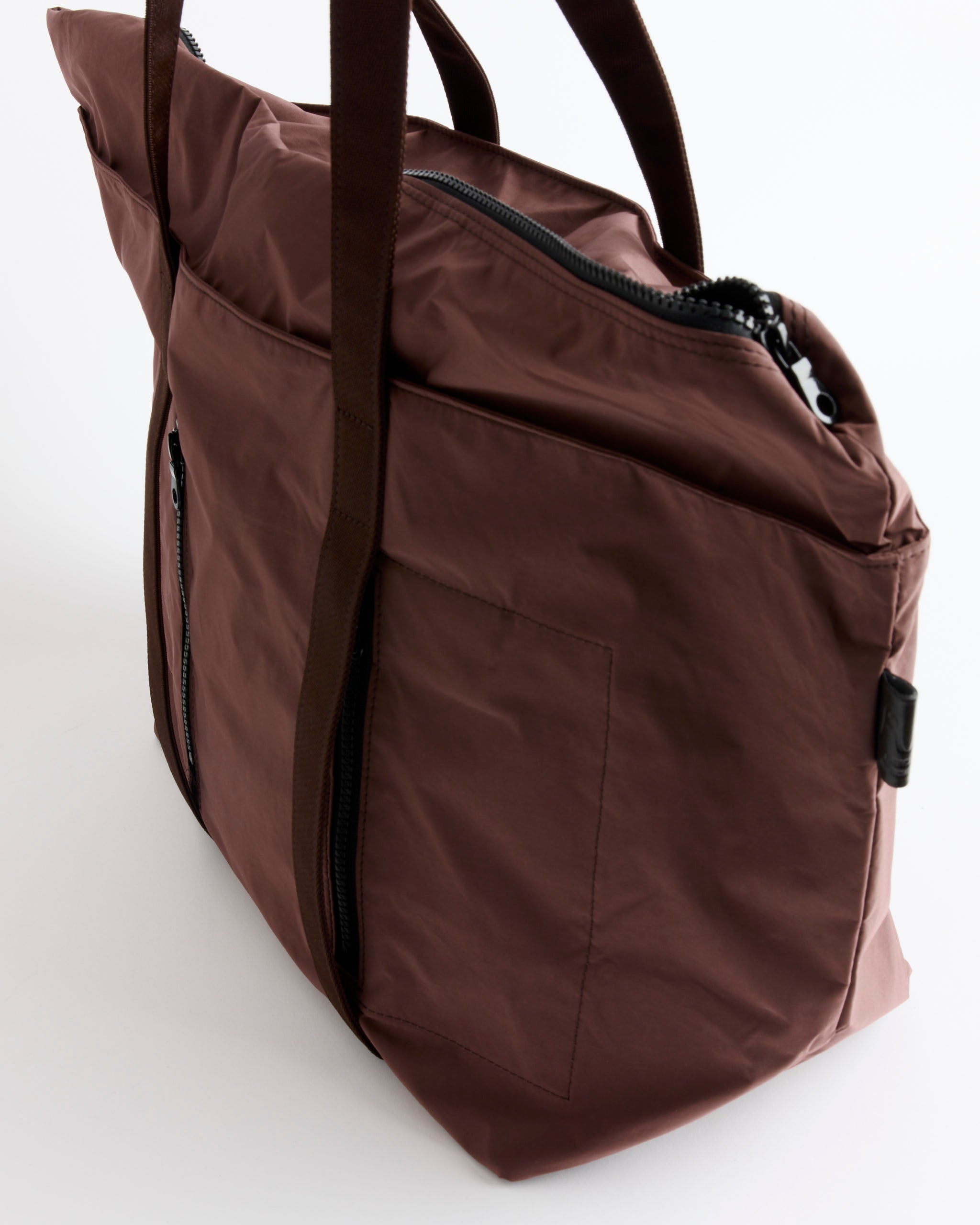 The Amiacalva Gabardine Tote in Bordeaux Brown, featuring black straps, a front zippered pocket, and a top zipper closure, is displayed against a white background.