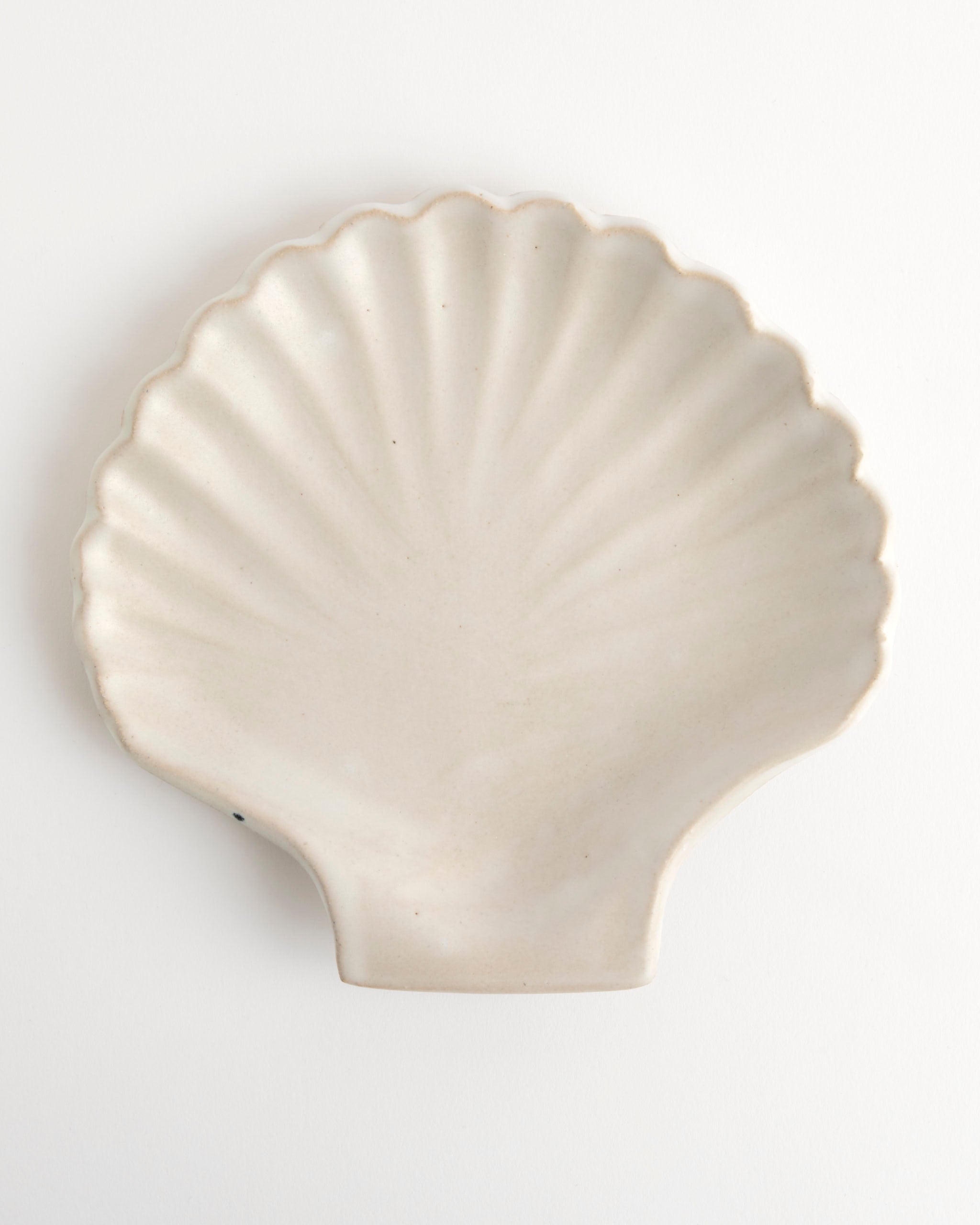 The Shell Plate by Beau Rush Ceramics features a smooth off-white surface with subtle ridges fanning out in a scallop shell shape, and is displayed on a white background.
