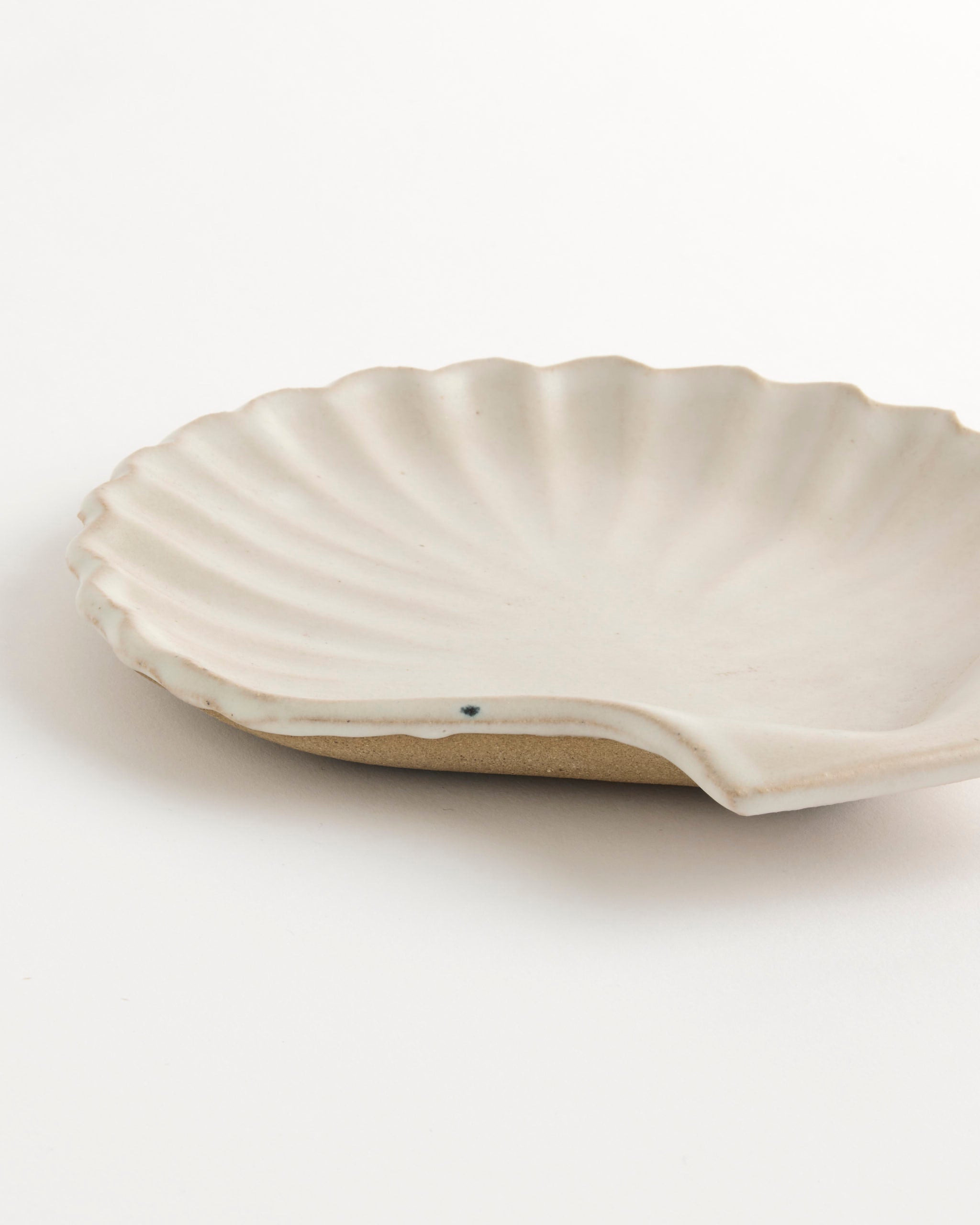 The Shell Plate by Beau Rush Ceramics features a wavy, textured edge and a matte off-white finish, designed in the shape of a scallop shell and displayed on a white surface.