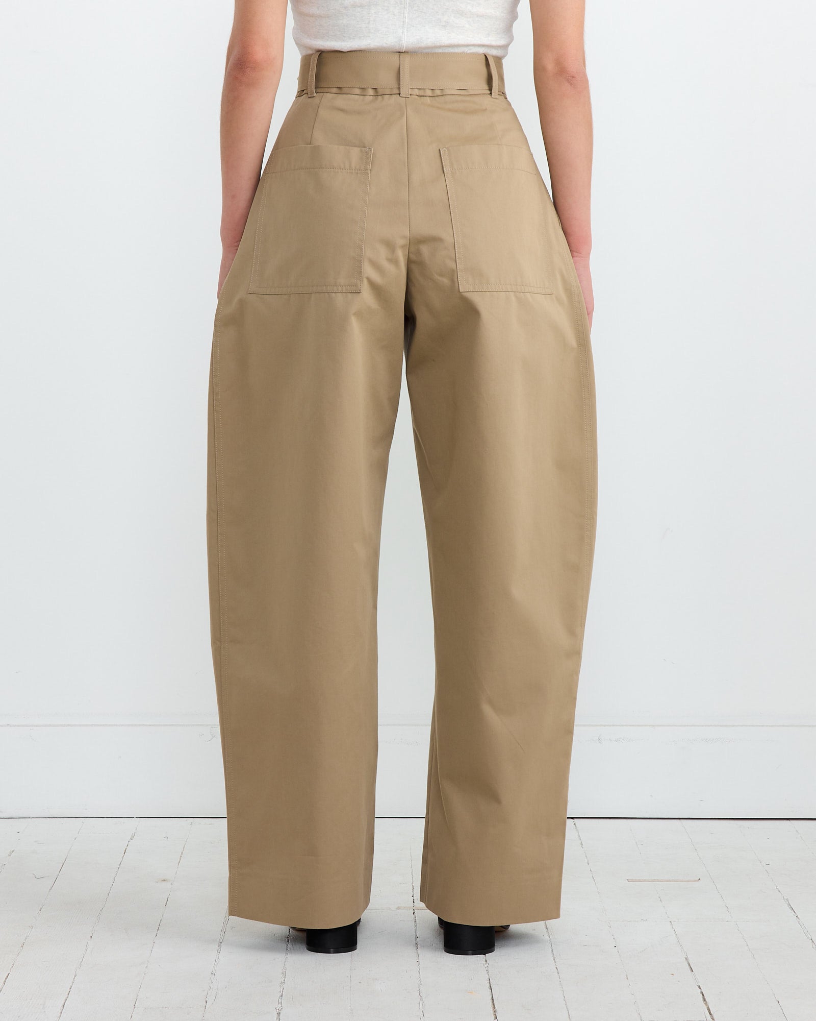 A rear view of someone in Studio Nicholsons Doring Pant in Parcel shows a contemporary silhouette with high-waisted, wide-leg khaki trousers paired with a white top, set against a seamless white backdrop. Their hands rest naturally at their sides on the crisp white floor.