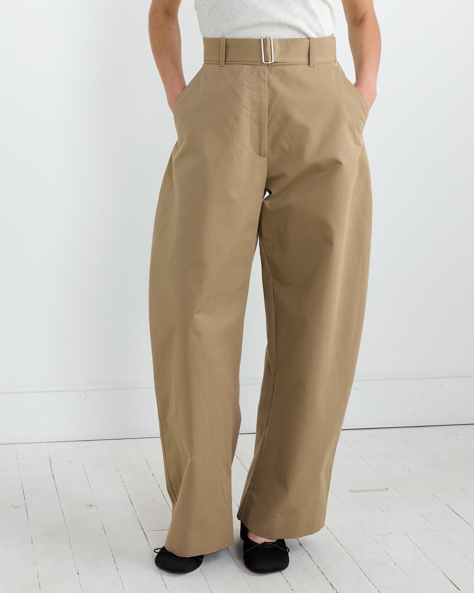 On a white wooden floor, a person embodies a contemporary silhouette in Studio Nicholsons Doring Pant in Parcel—tan wide-leg trousers made from a cotton CORDURA® nylon-blend with a belt. Hands are in pockets, black shoes complete the look against the plain white backdrop.