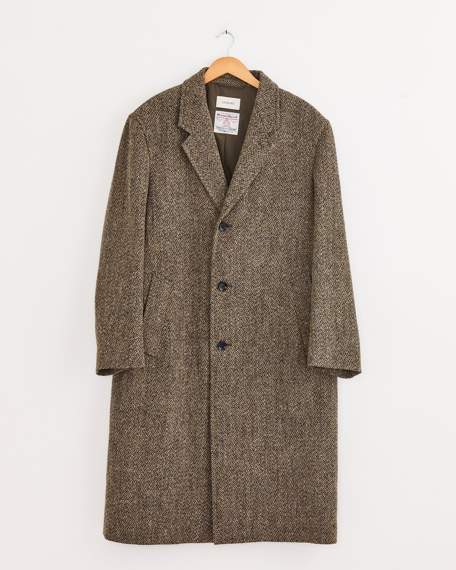 A Lemaire Chesterfield Coat in Black/Straw Melange with a notched collar and three black buttons hangs on a wooden hanger against a plain white wall.