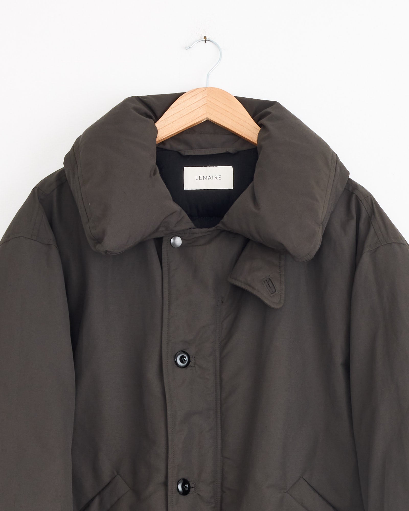 The Puffer Blouson in Dark Espresso by Lemaire hangs on a wooden hanger, featuring a large collar and black buttons. A label inside the collar displays the brand name, set against a plain white background.