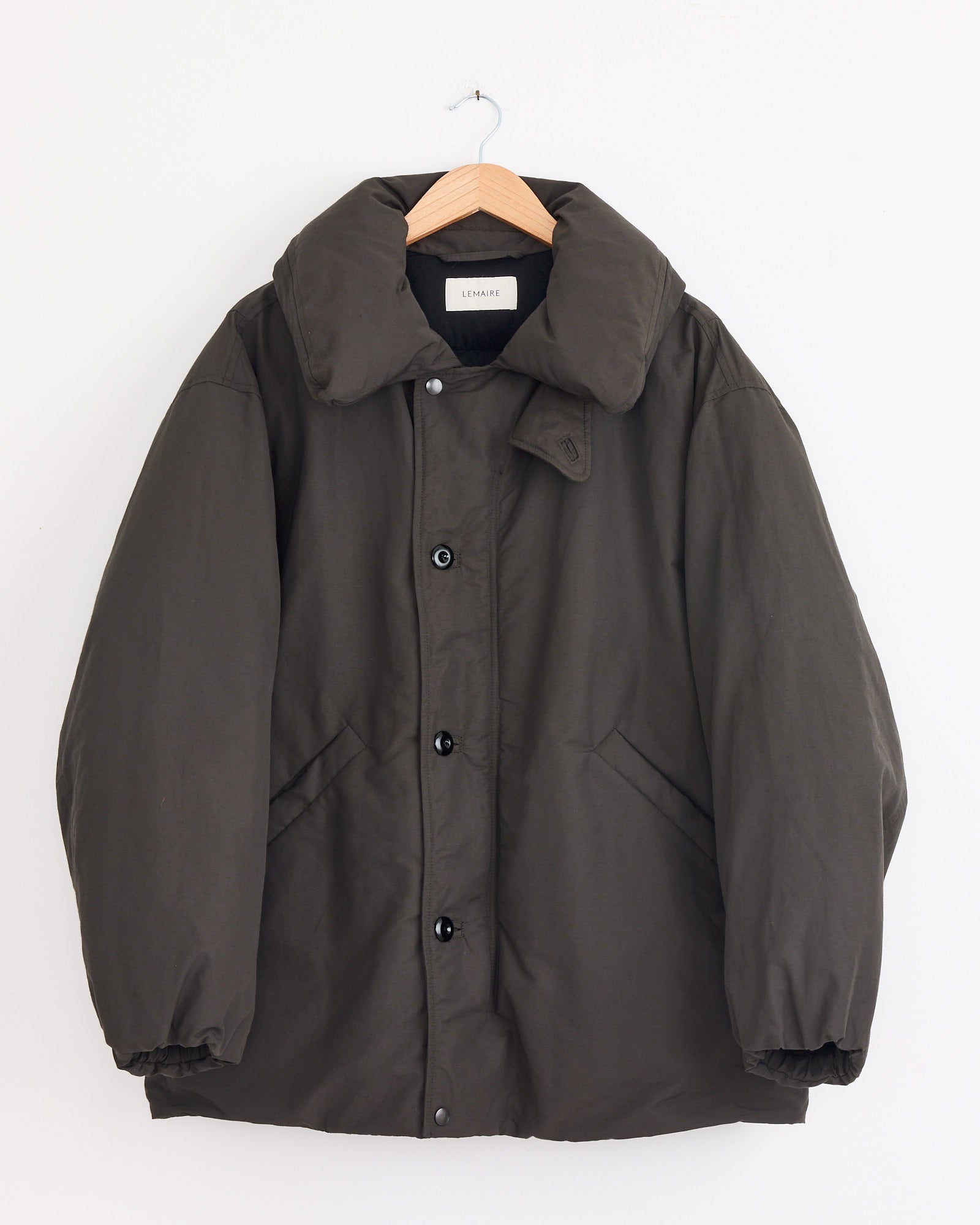 A Lemaire Puffer Blouson in Dark Espresso, featuring a large collar, front buttons, and elastic cuffs, hangs on a wooden hanger against a plain white wall.