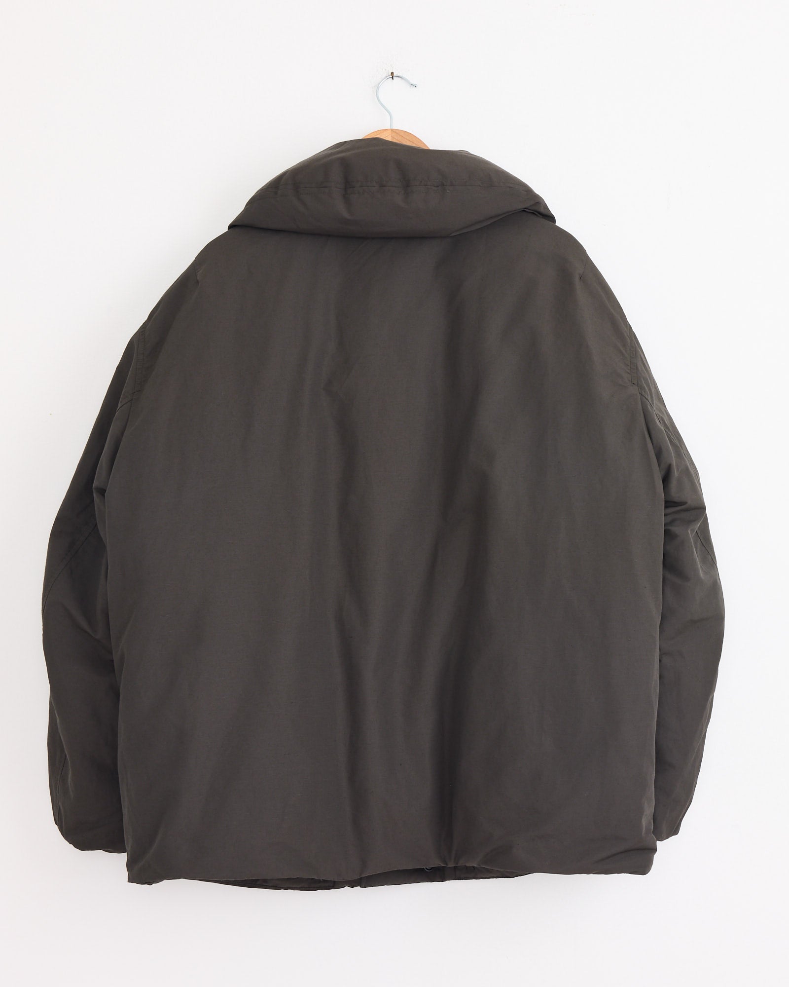 A Lemaire Puffer Blouson in Dark Espresso hangs on a wooden hanger against a plain white wall, displayed from the back.