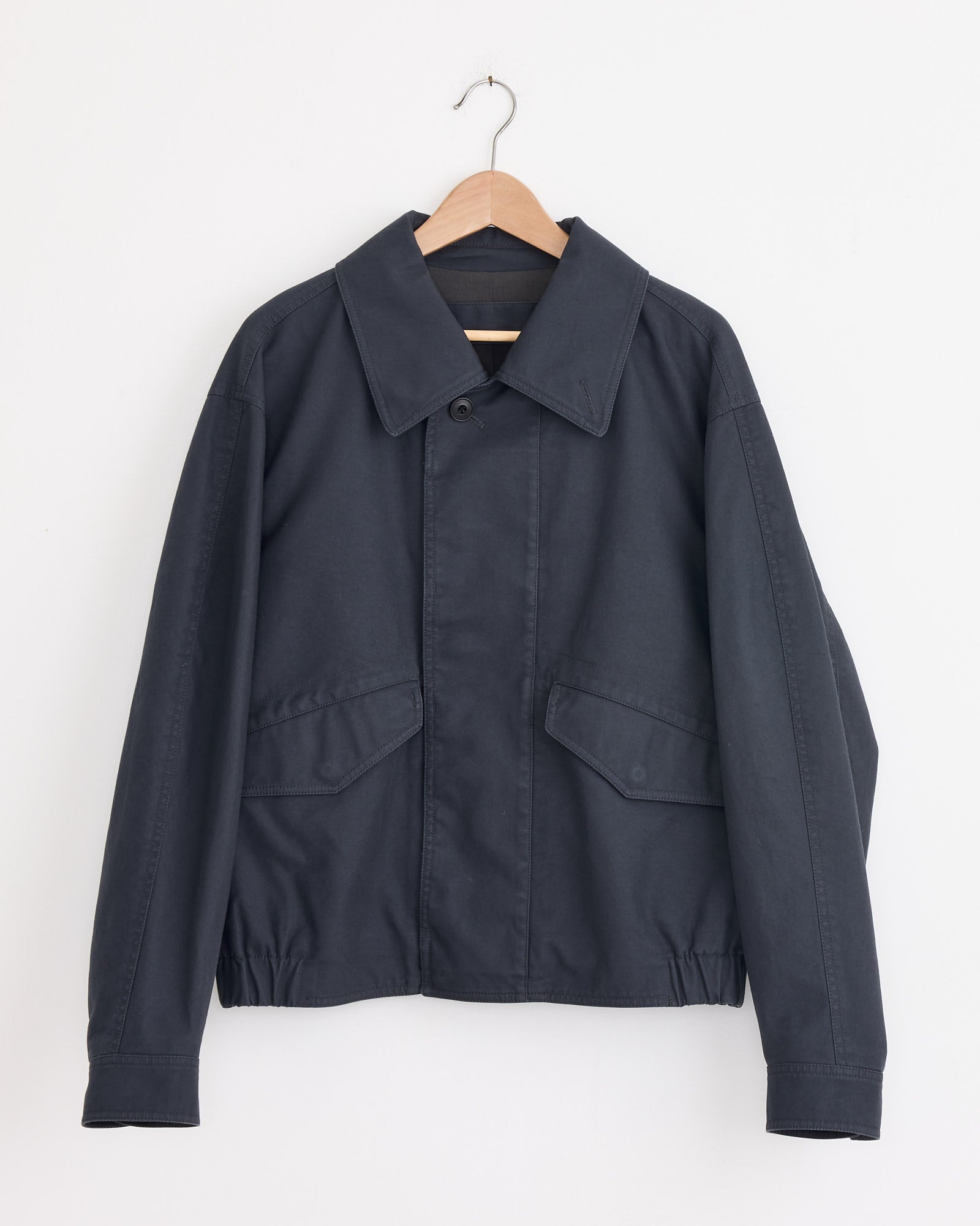 The Lemaire Boxy Blouson in Graphite Blue is a cotton gabardine, long-sleeve jacket with a collar, button closure, two front flap pockets, and a straight hem, shown on a wooden hanger against a white wall.