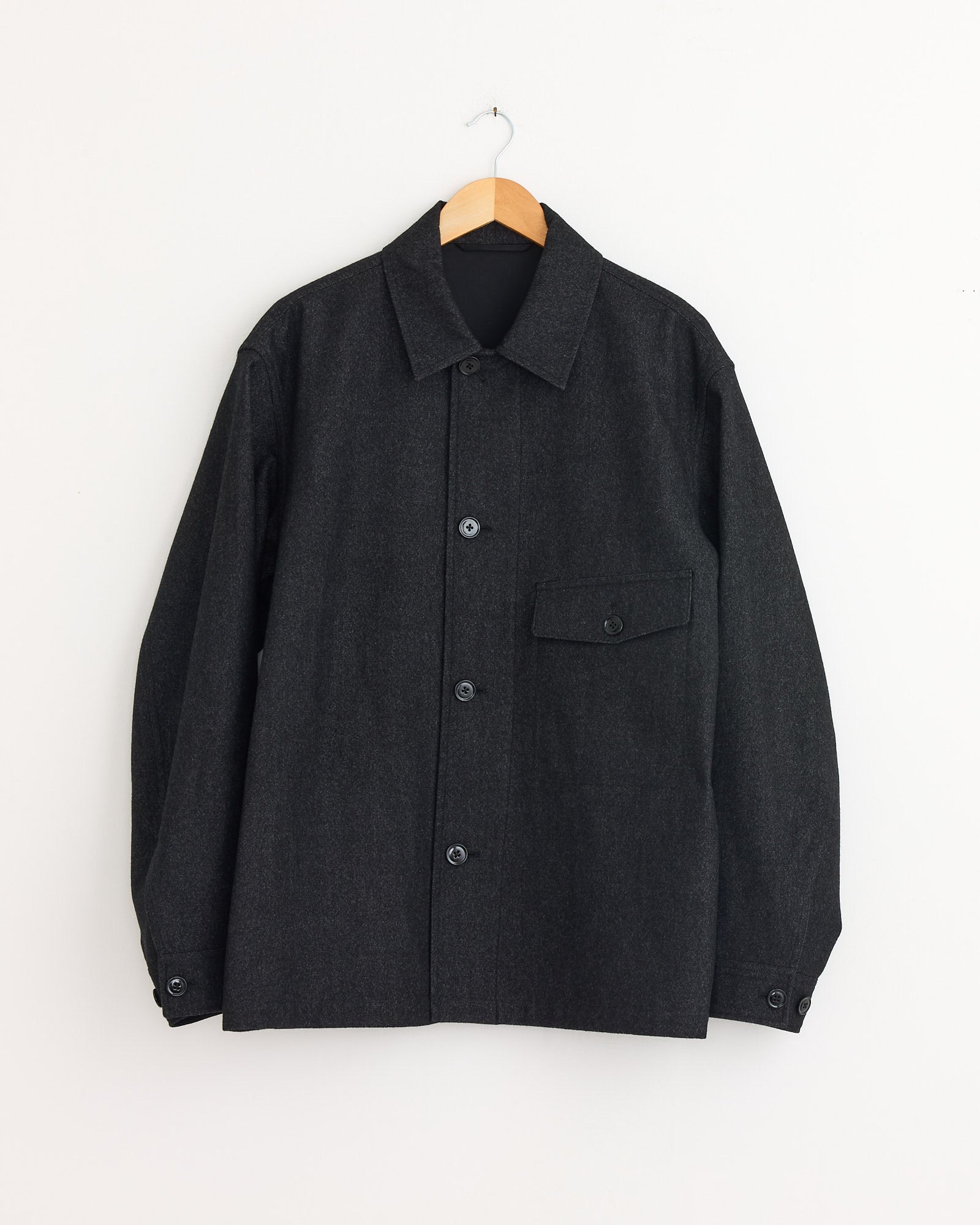 The Lemaire One Pocket Overshirt in Black/Grey Melange, featuring a boxy fit, long sleeves, collar, and a single flap chest pocket, hangs on a wooden hanger against a white wall.