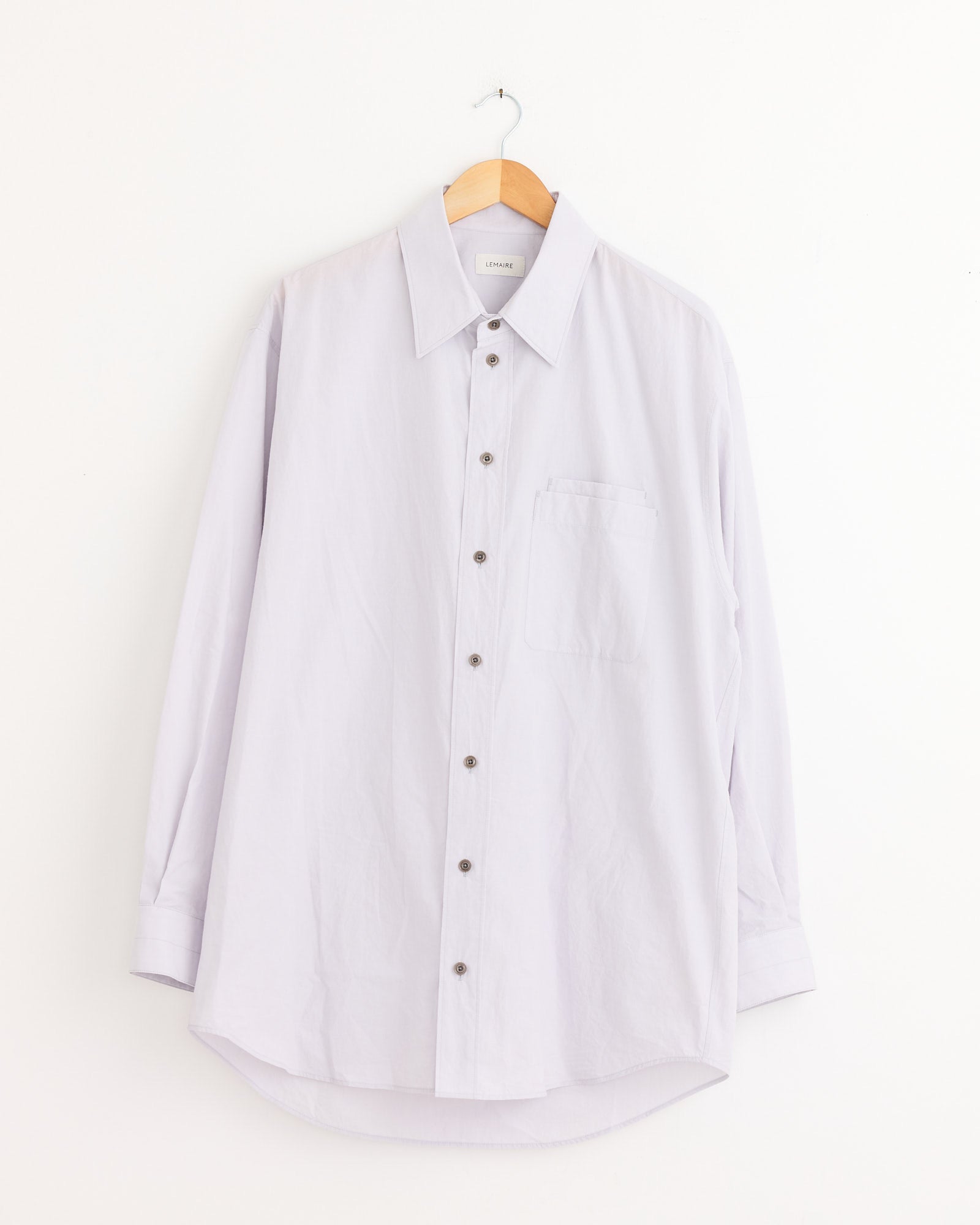 The Lemaire Double Pocket Shirt in Misty Lilac, made of cotton crepe, with long sleeves and double front pockets, hangs on a wooden hanger against a plain white wall.