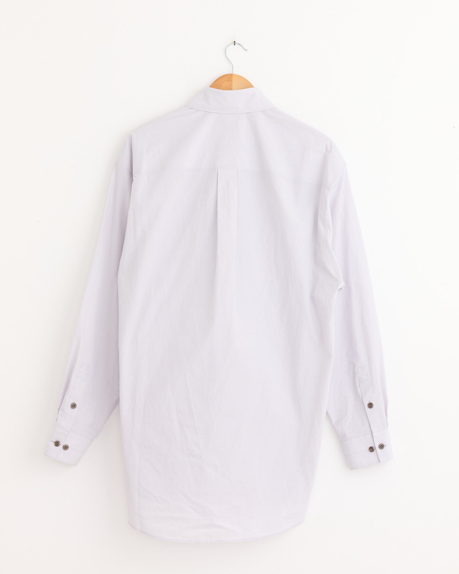 A Lemaire Double Pocket Shirt in Misty Lilac, featuring long sleeves, hangs on a wooden hanger against a white wall and is shown from the back.