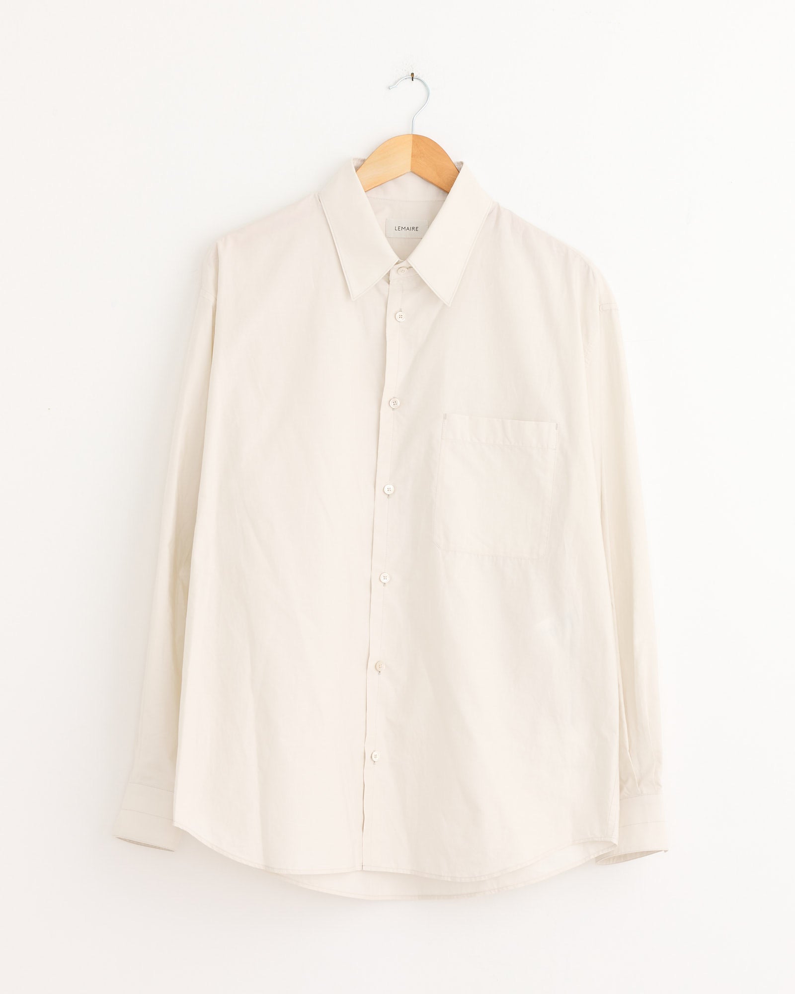 The Lemaire Regular Shirt in Light Cream is a long-sleeved, collared button-up with a chest pocket, displayed on a wooden hanger against a white background. For sizing or fit questions, our online chat can help.