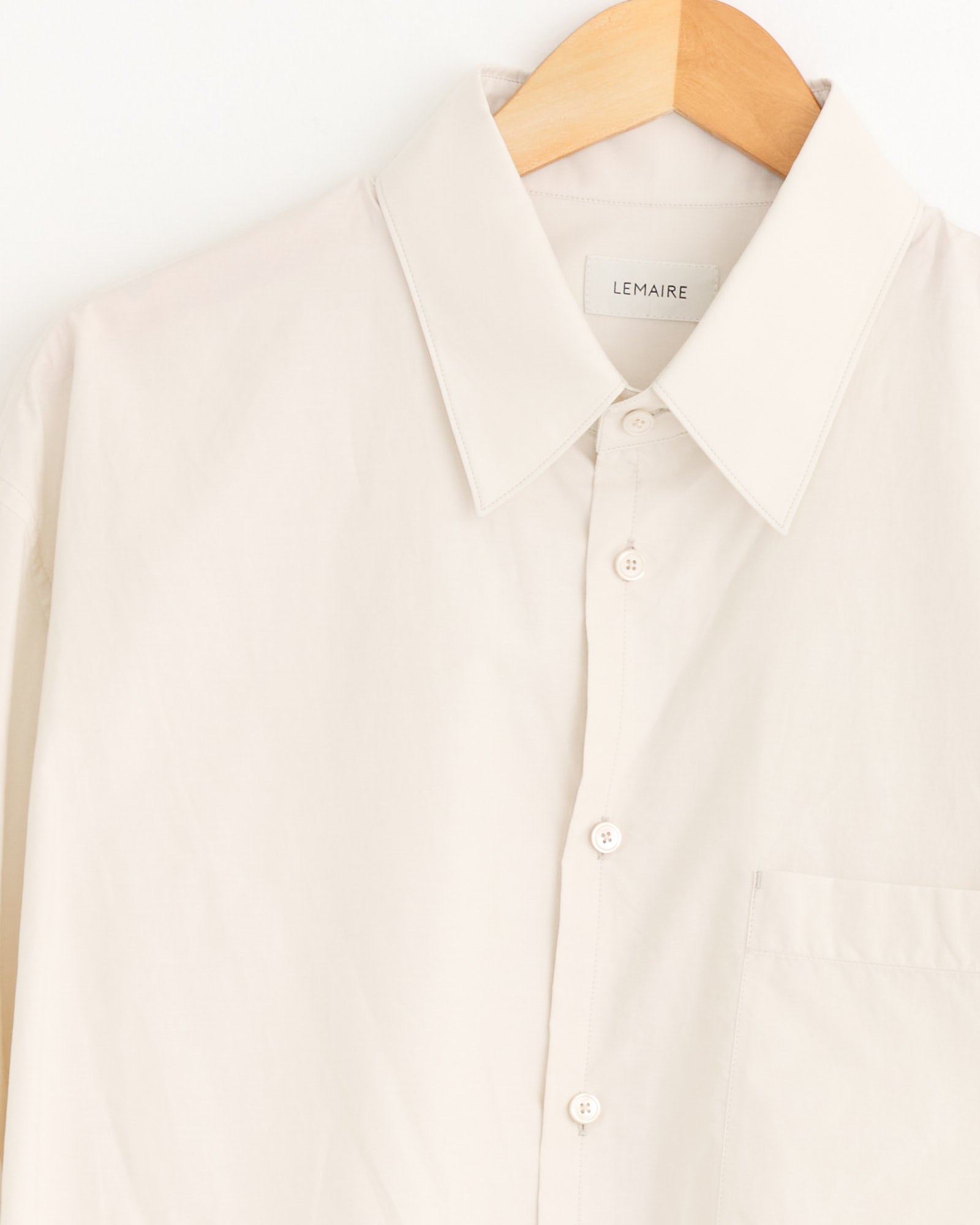 The Regular Shirt in Light Cream by Lemaire features a chest pocket, pointed collar, and visible label. It hangs on a wooden hanger. For sizing or fit questions, please use our online chat for assistance.