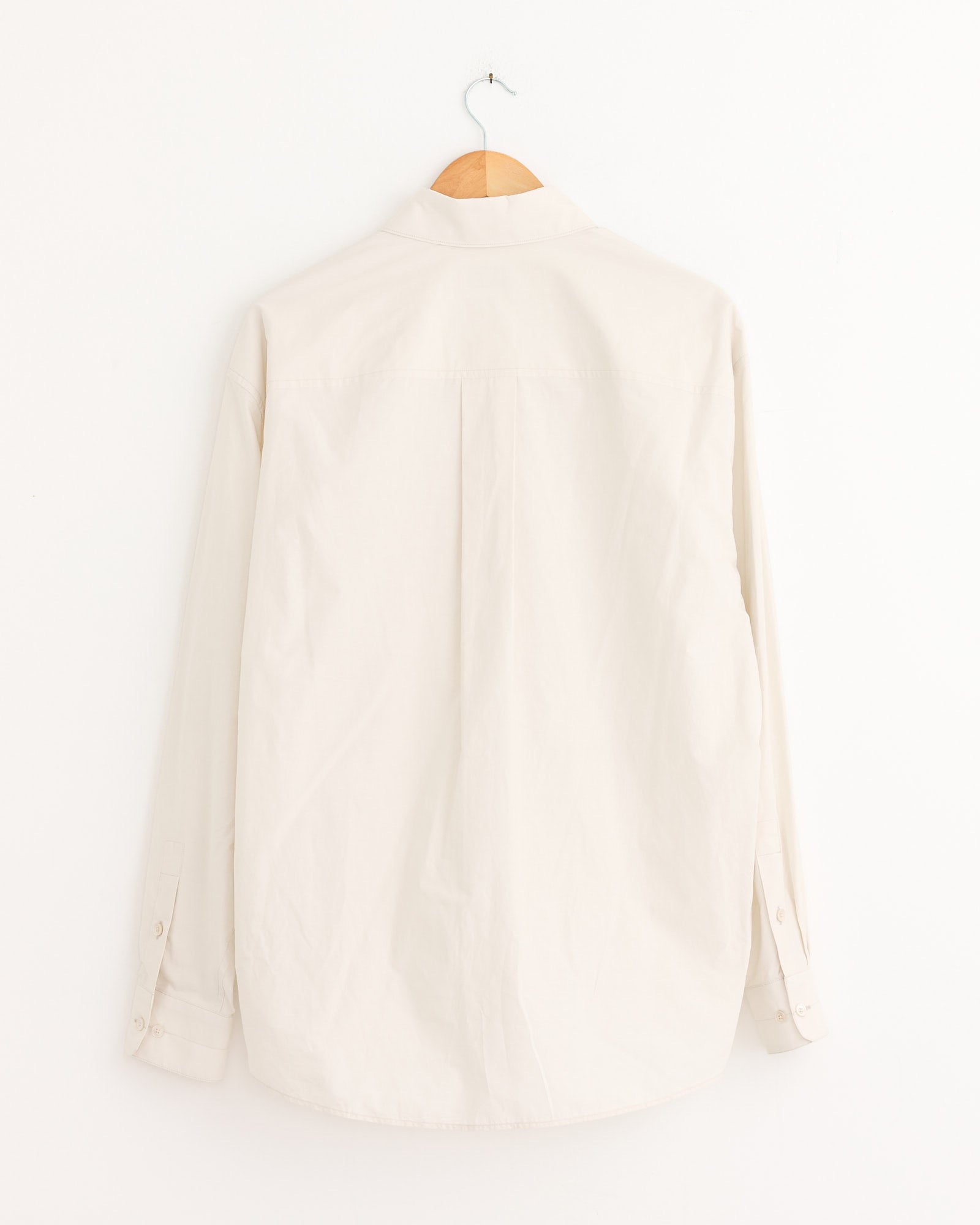 The Lemaire Regular Shirt in Light Cream, shown from the back on a wooden hanger against a white wall, features long sleeves and a button-up design. For fit or sizing questions, our online chat is available to help you.