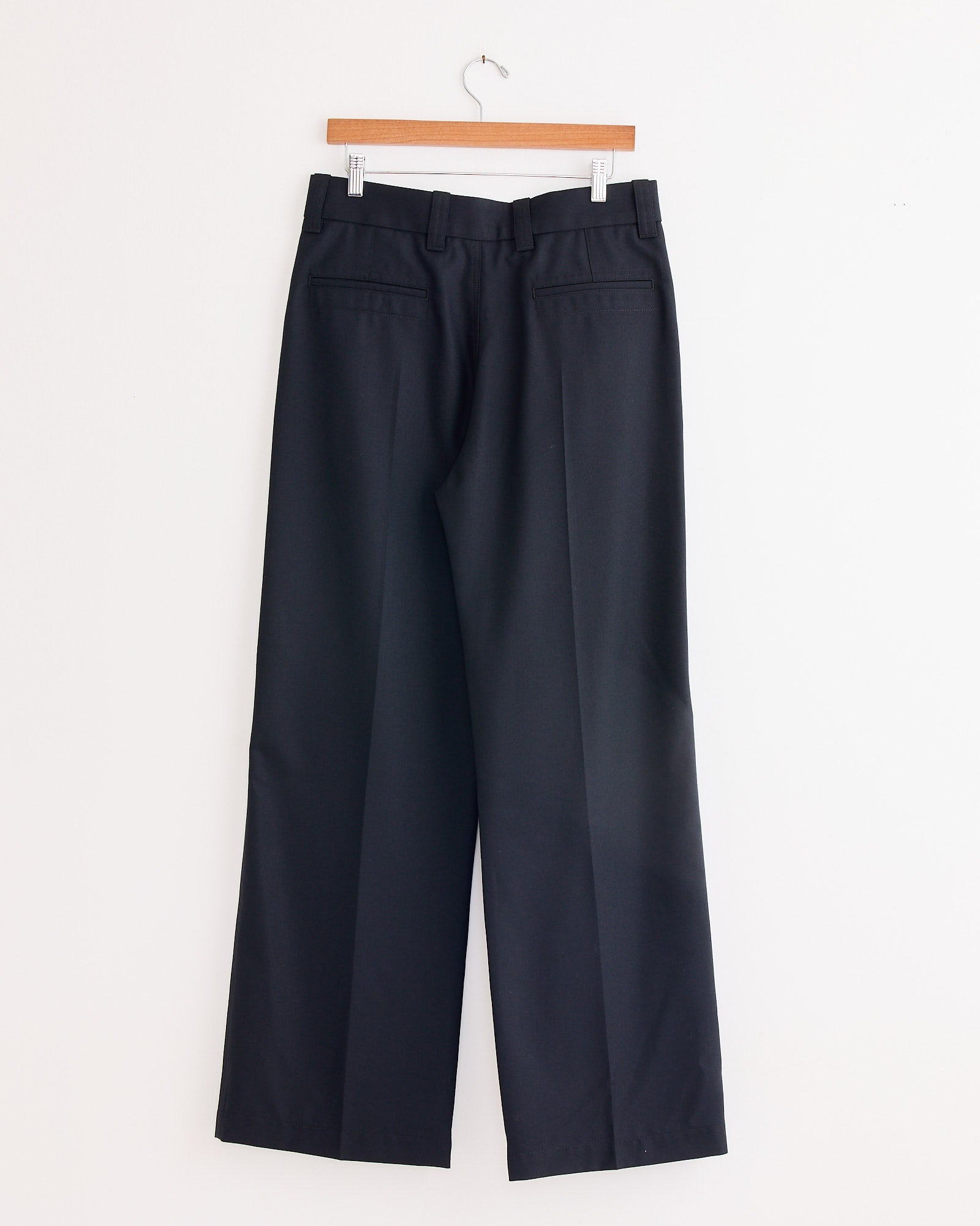 The Straight Tailored Pant in Marine Melange by Lemaire, featuring belt loops and back welt pockets, hangs on a wooden hanger against a white wall—highlighting classic, traditional tailoring.