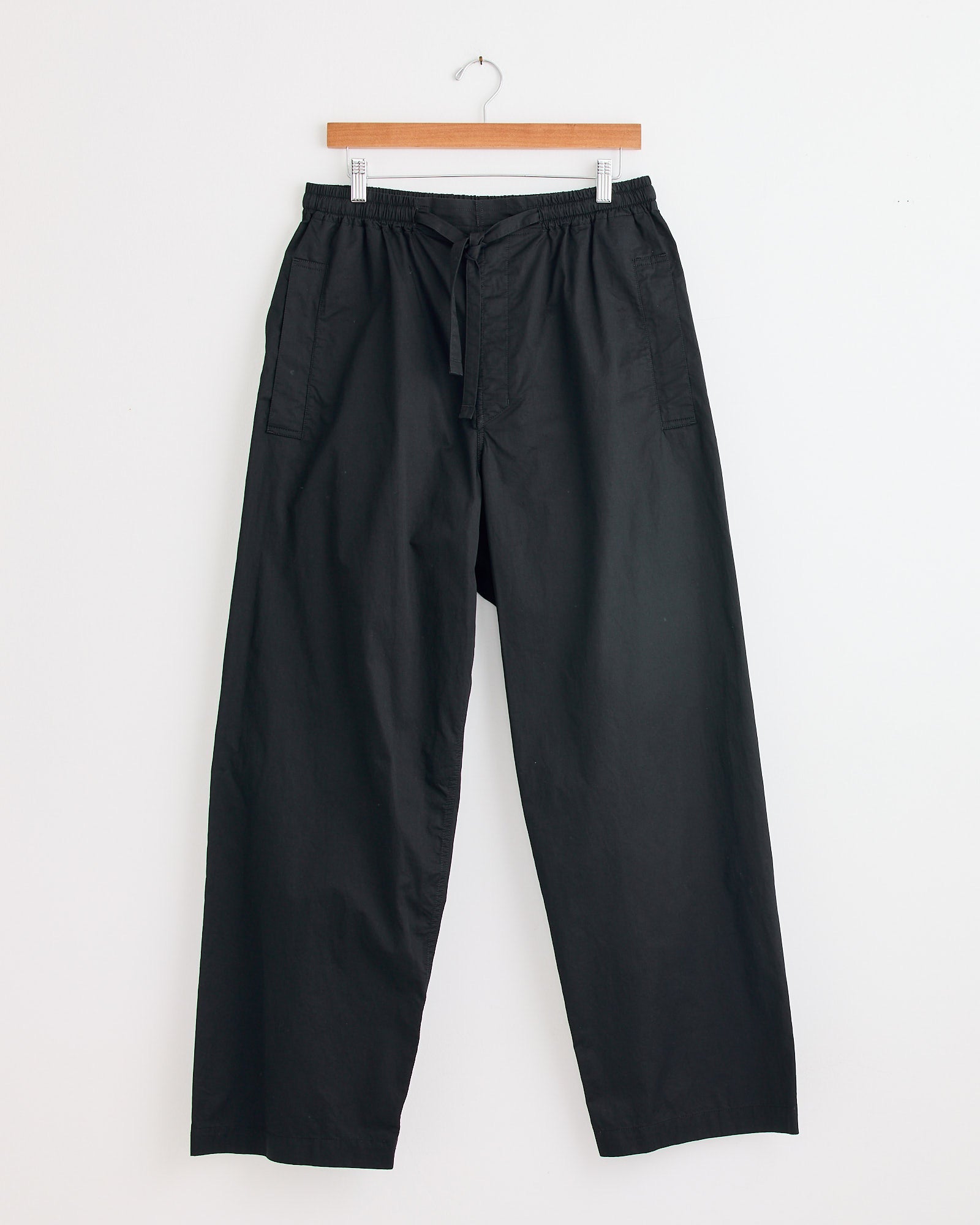 The Lemaire Pyjama Pant in Black features a loose fit, elastic waistband, drawstring, and side pockets. Displayed on a hanger against a white wall—contact our online chat for sizing help.