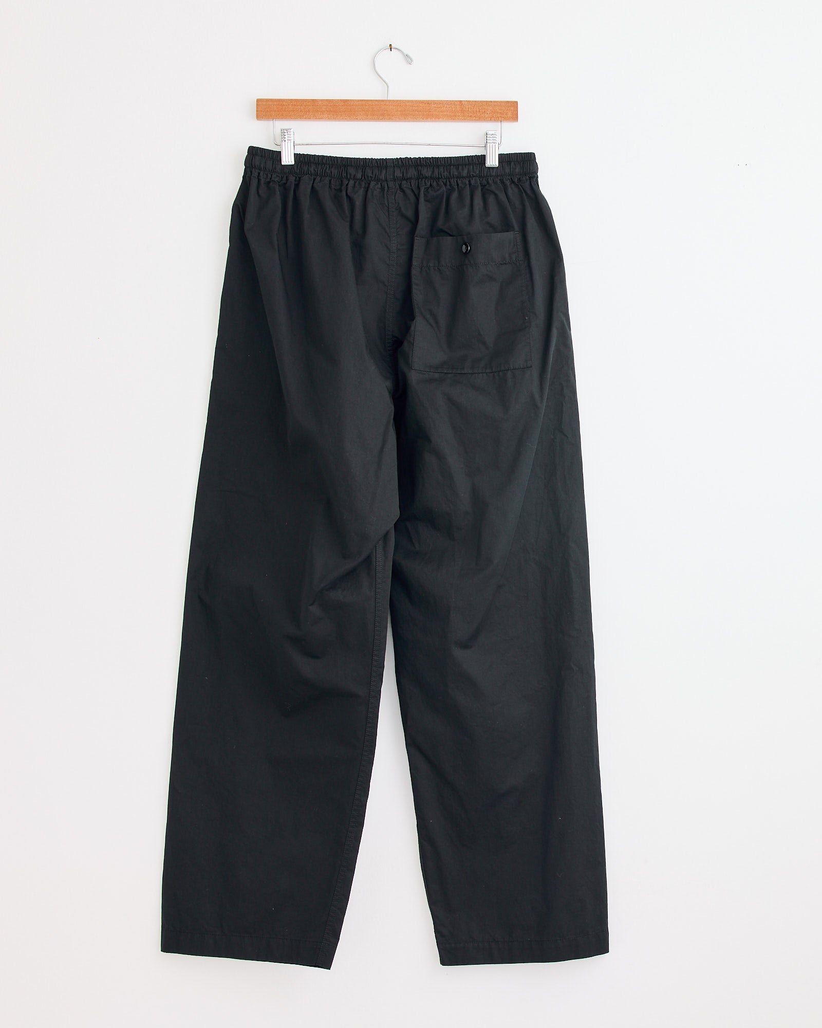 The Lemaire Pyjama Pant in Black features a relaxed fit, elastic waistband, and single back pocket. Shown on a wooden hanger against a white wall. Contact our online chat for sizing or fit questions before ordering.