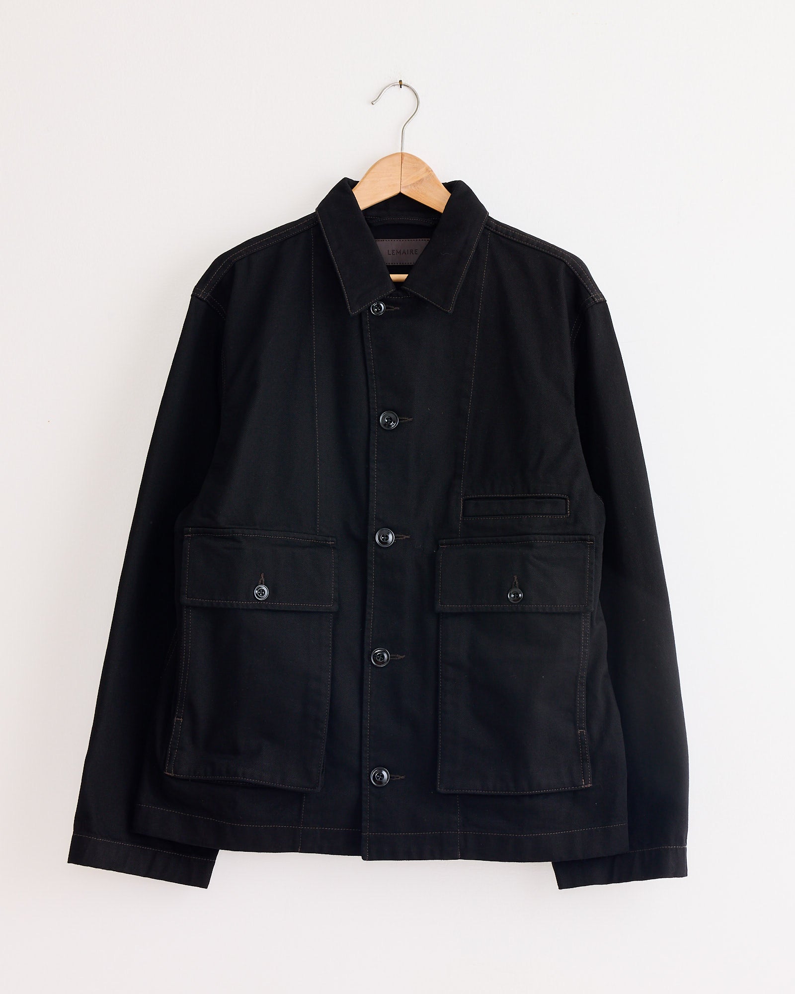 The Boxy Jacket in Black by Lemaire features large front pockets and a collar, shown here hanging on a wooden hanger against a plain white wall.