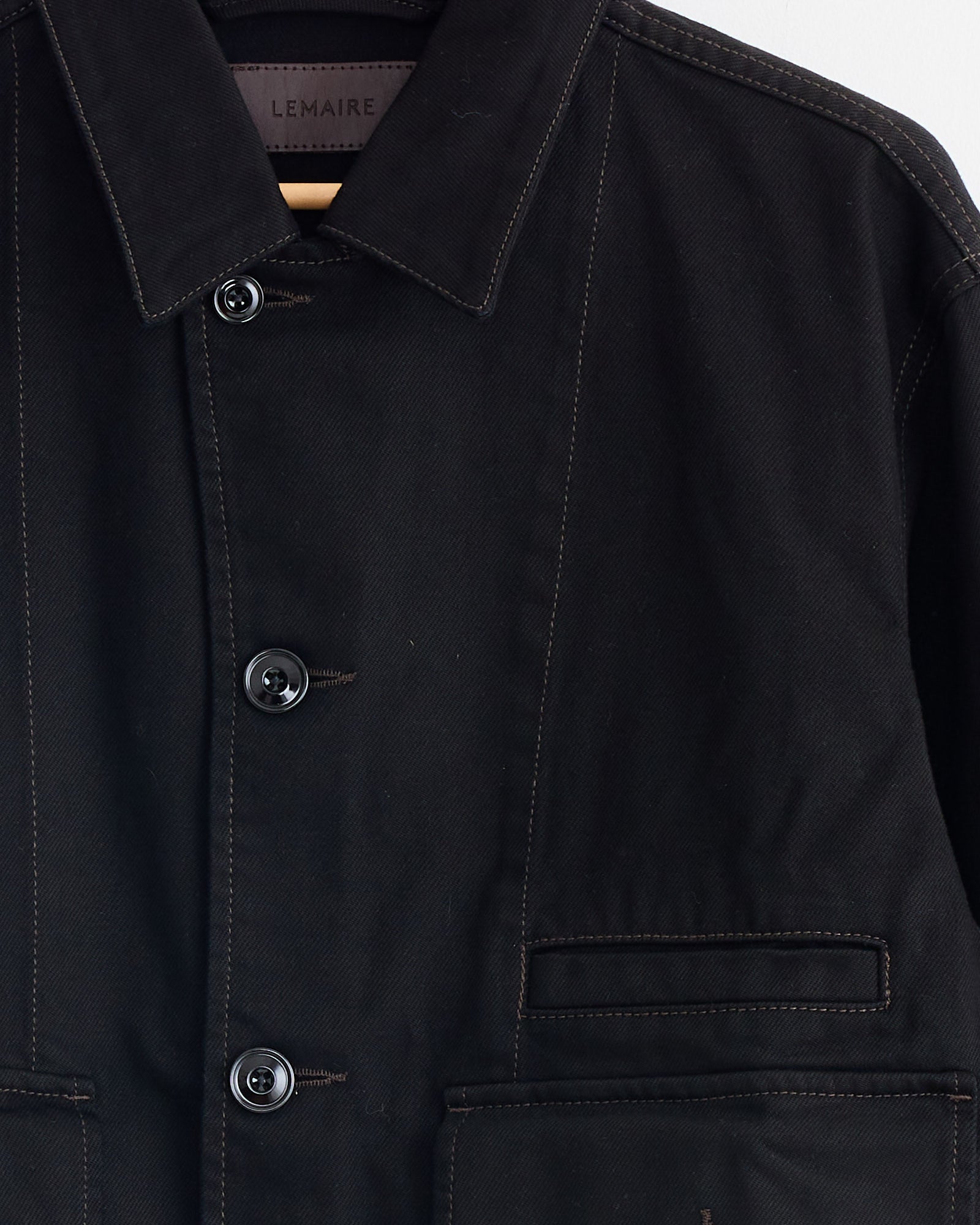 A close-up of the Lemaire Boxy Jacket in Black, showcasing visible stitching, two horizontal front pockets, and a label inside the collar with the Lemaire branding.