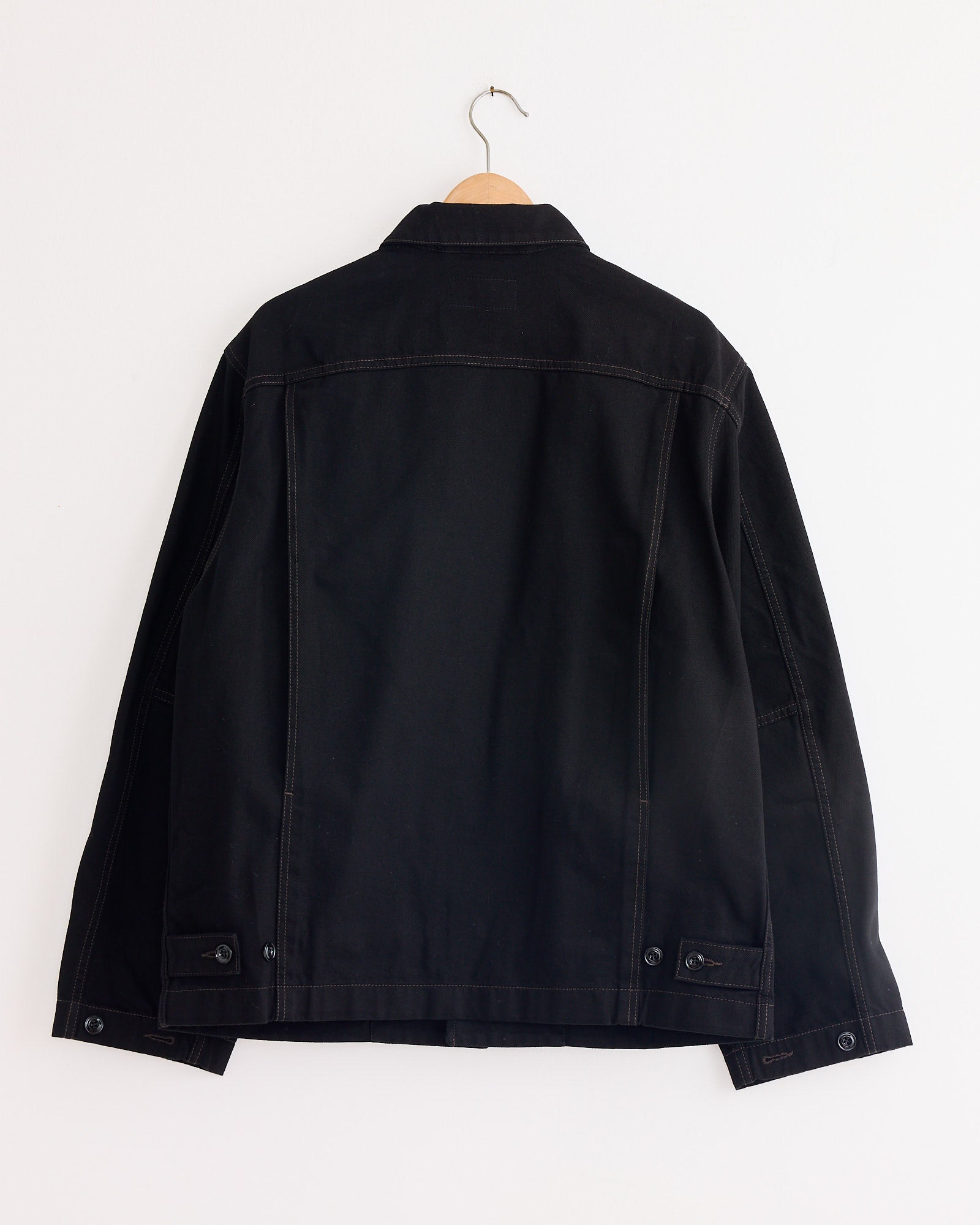 The Lemaire Boxy Jacket in Black hangs on a wooden hanger against a plain white wall, shown from the back to highlight its simple stitching and classic, boxy silhouette—a timeless unisex piece for any wardrobe.