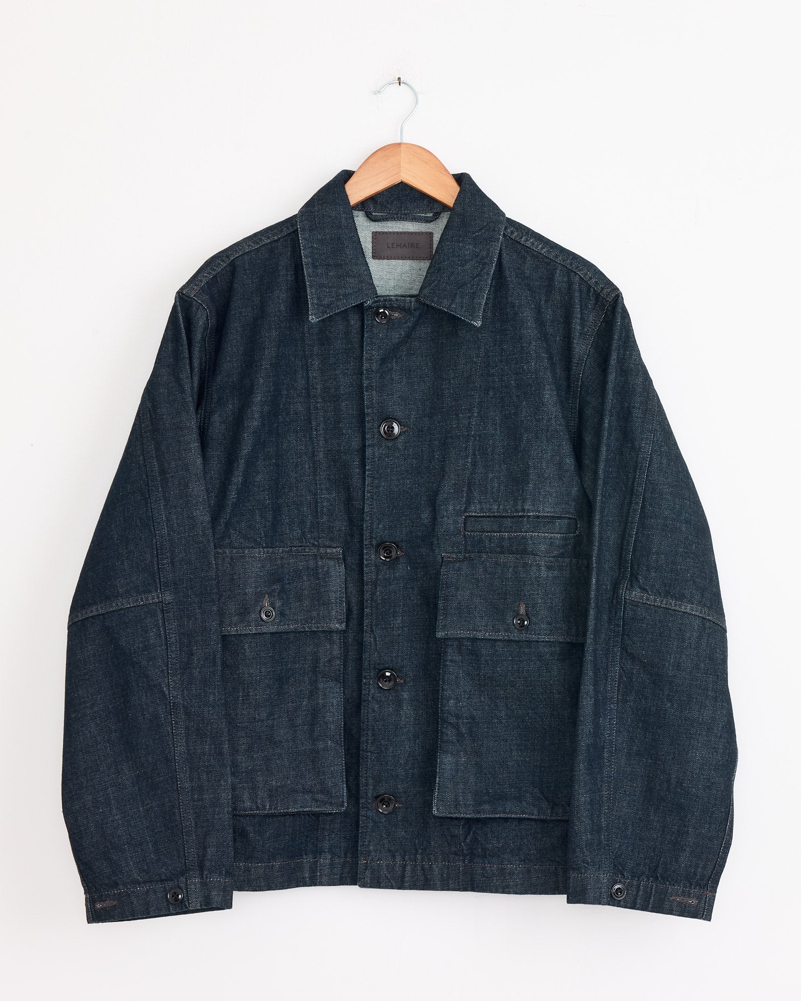 The Lemaire Boxy Jacket in Denim Indigo features black buttons, a pointed collar, and two large front pockets. It is shown hanging on a wooden hanger against a white background.