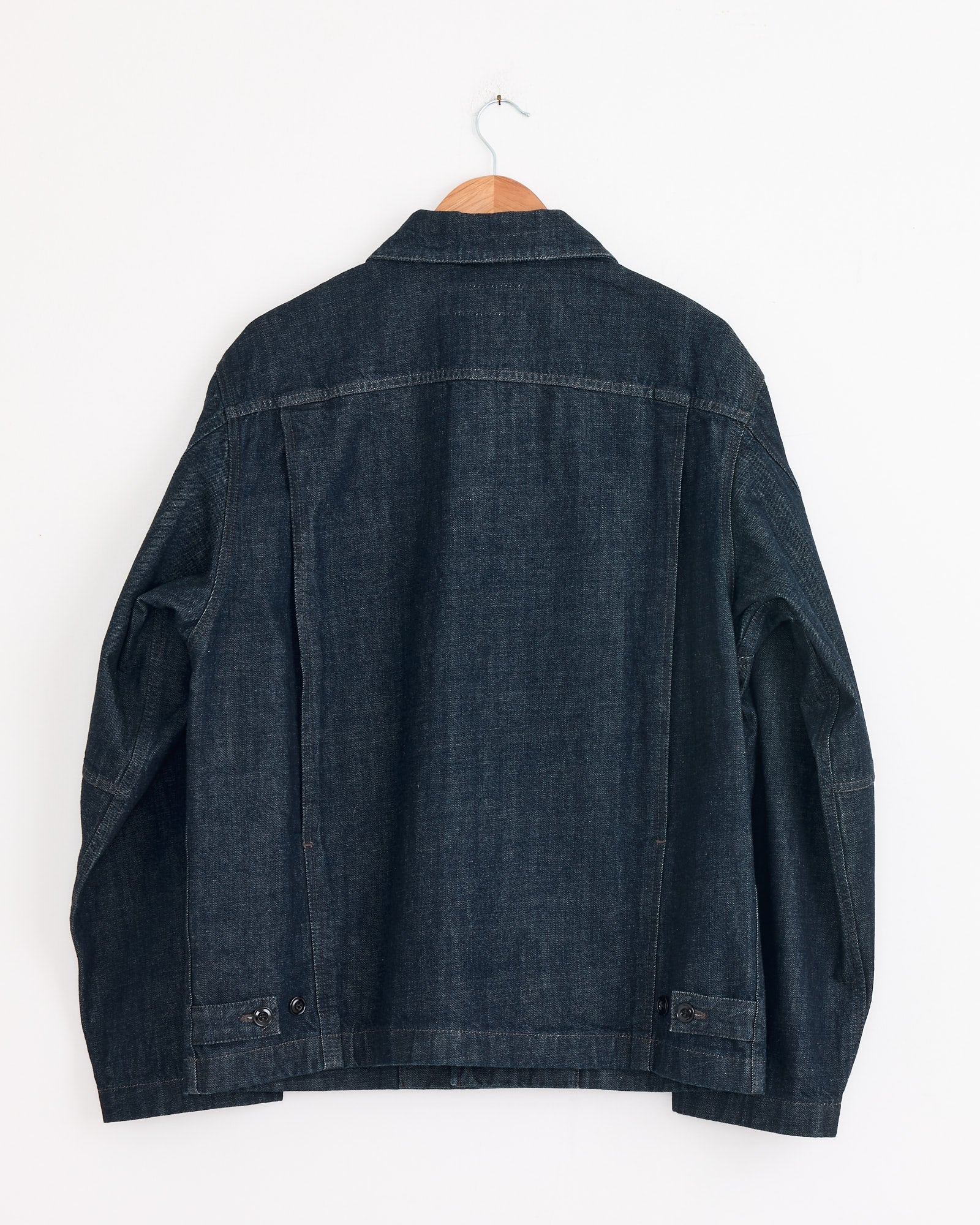 The Lemaire Boxy Jacket in Denim Indigo hangs on a wooden hanger against a plain white wall, shown from the back.