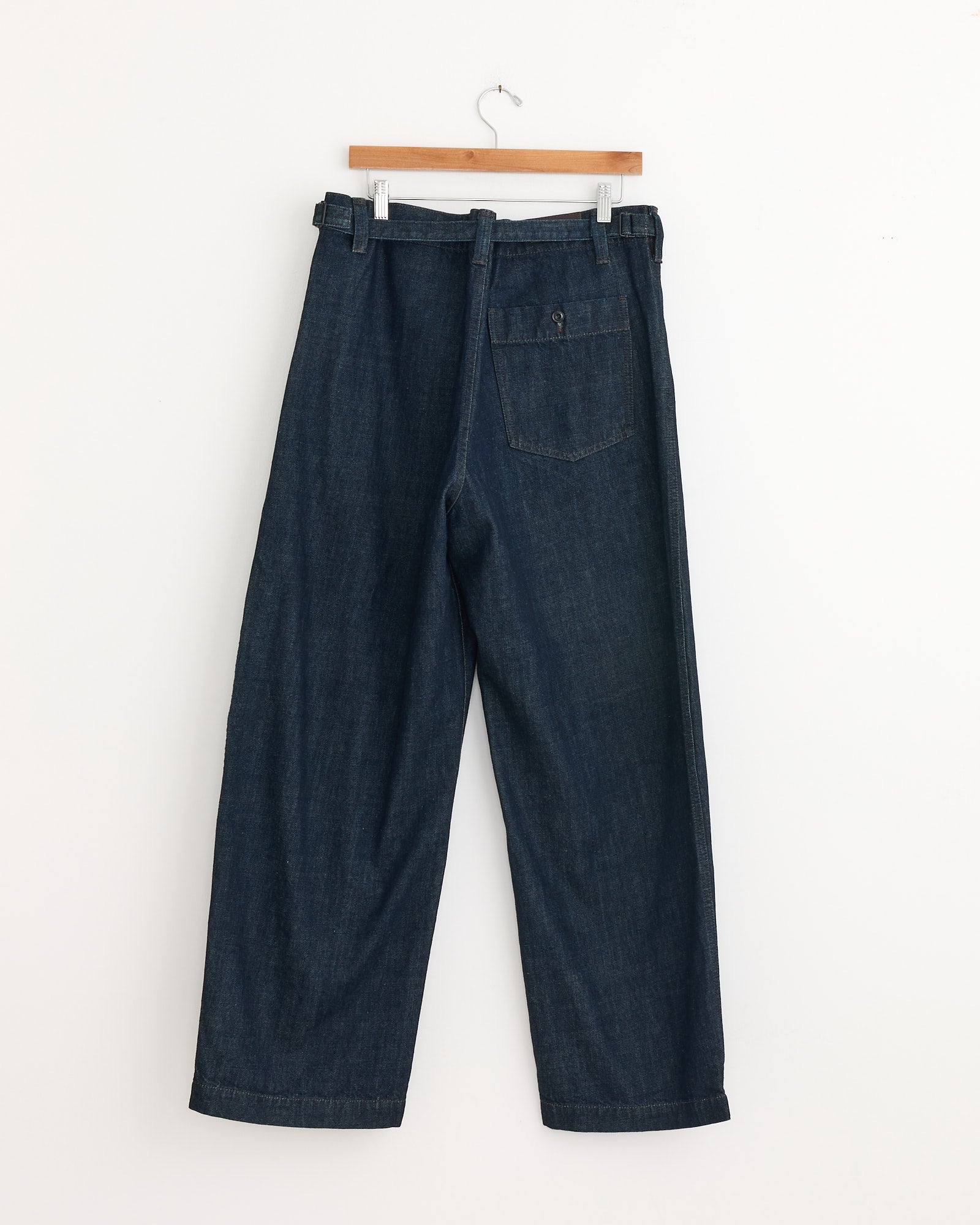 The Lemaire Maxi Workwear Pant in Denim Indigo features thick denim, a loose straight-leg fit, a right-side back pocket, and a matching belt, shown hanging on a wooden hanger against a white wall.