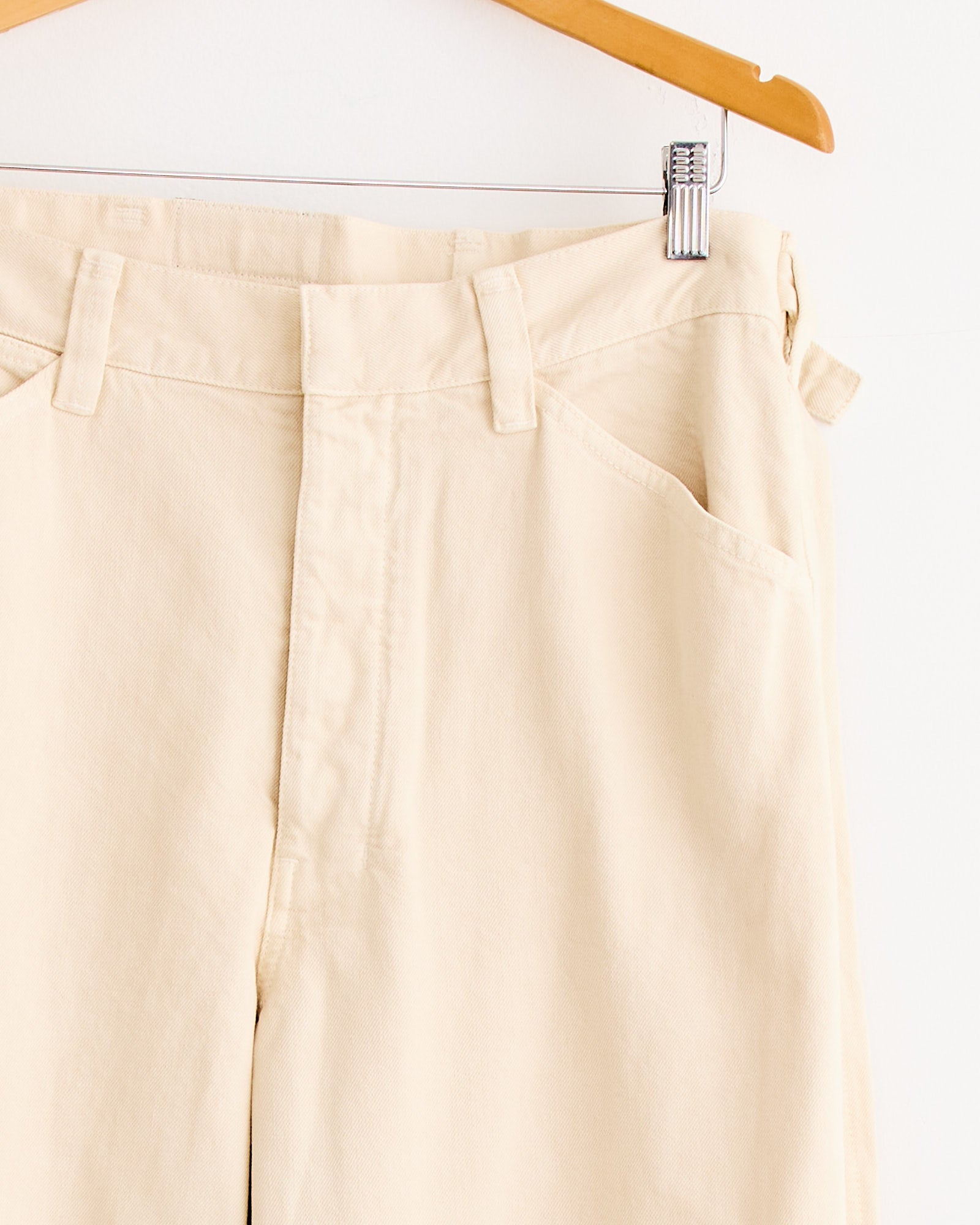 Close-up of Lemaire Maxi Workwear Pant in Denim Snow Clay on a wooden hanger, showcasing the waistband, belt loops, pockets, and upper legs in classic Lemaire style against a plain white background.