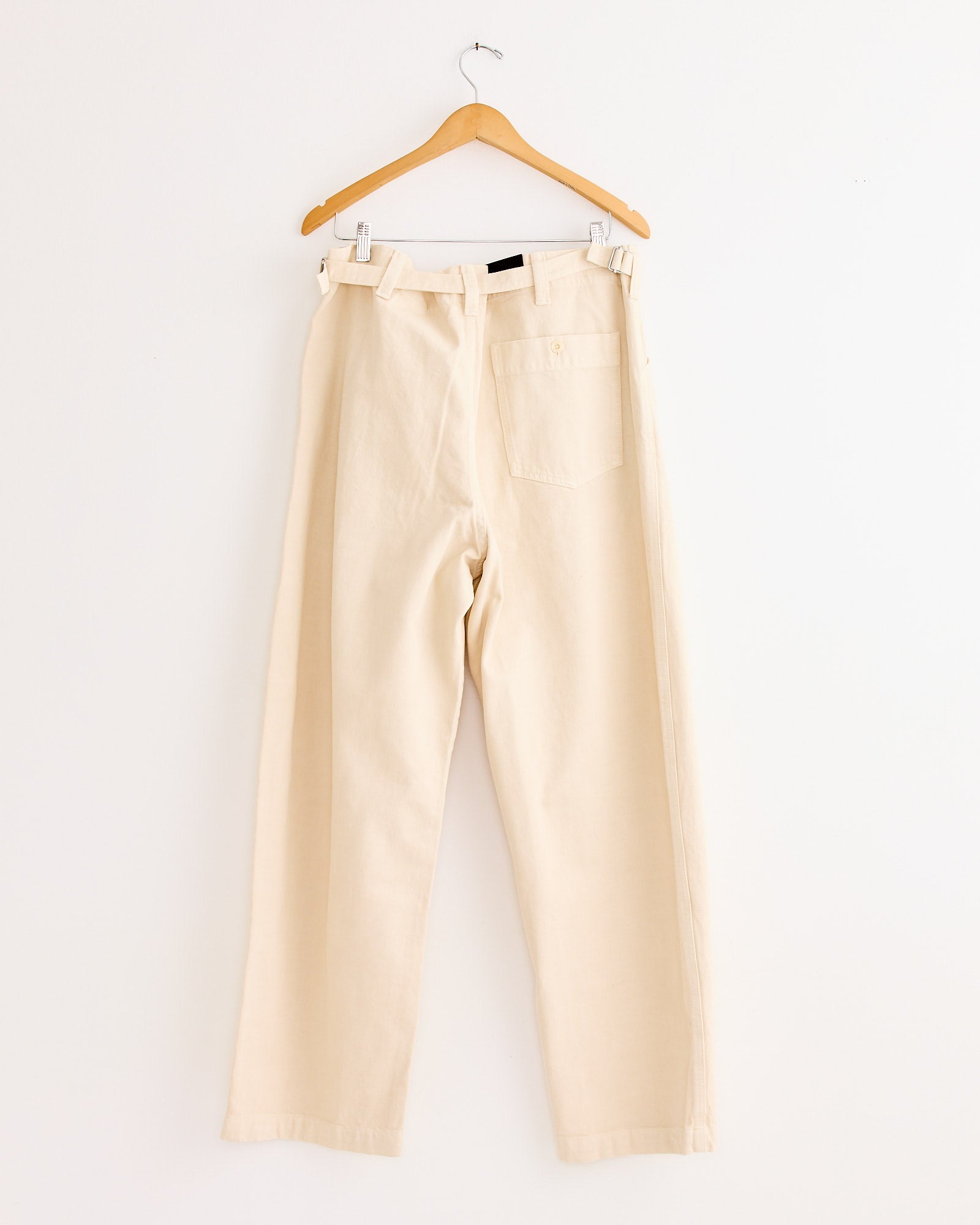 The Maxi Workwear Pant in Denim Snow Clay by Lemaire—high-waisted, cream-toned, straight-leg with belt loops and a single back pocket—hangs on a wooden hanger against a white wall, showcasing the brand’s minimalist style.
