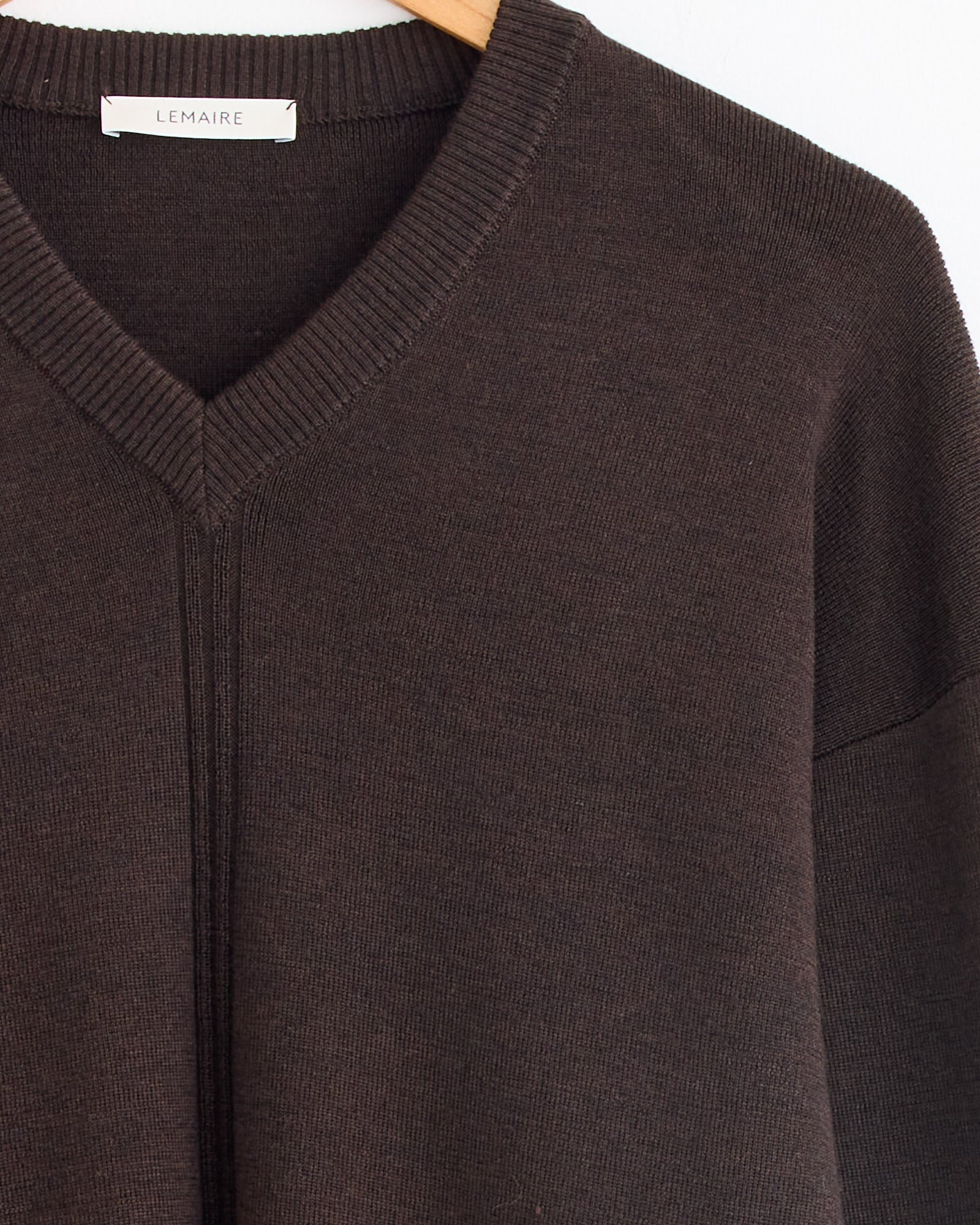 Close-up of the Lemaire V Neck Sweater in Dark Cacao on a hanger, showing ribbed neckline detailing, a central knit line, and the white LEMAIRE label. For sizing or fit questions, our online chat is available to assist.