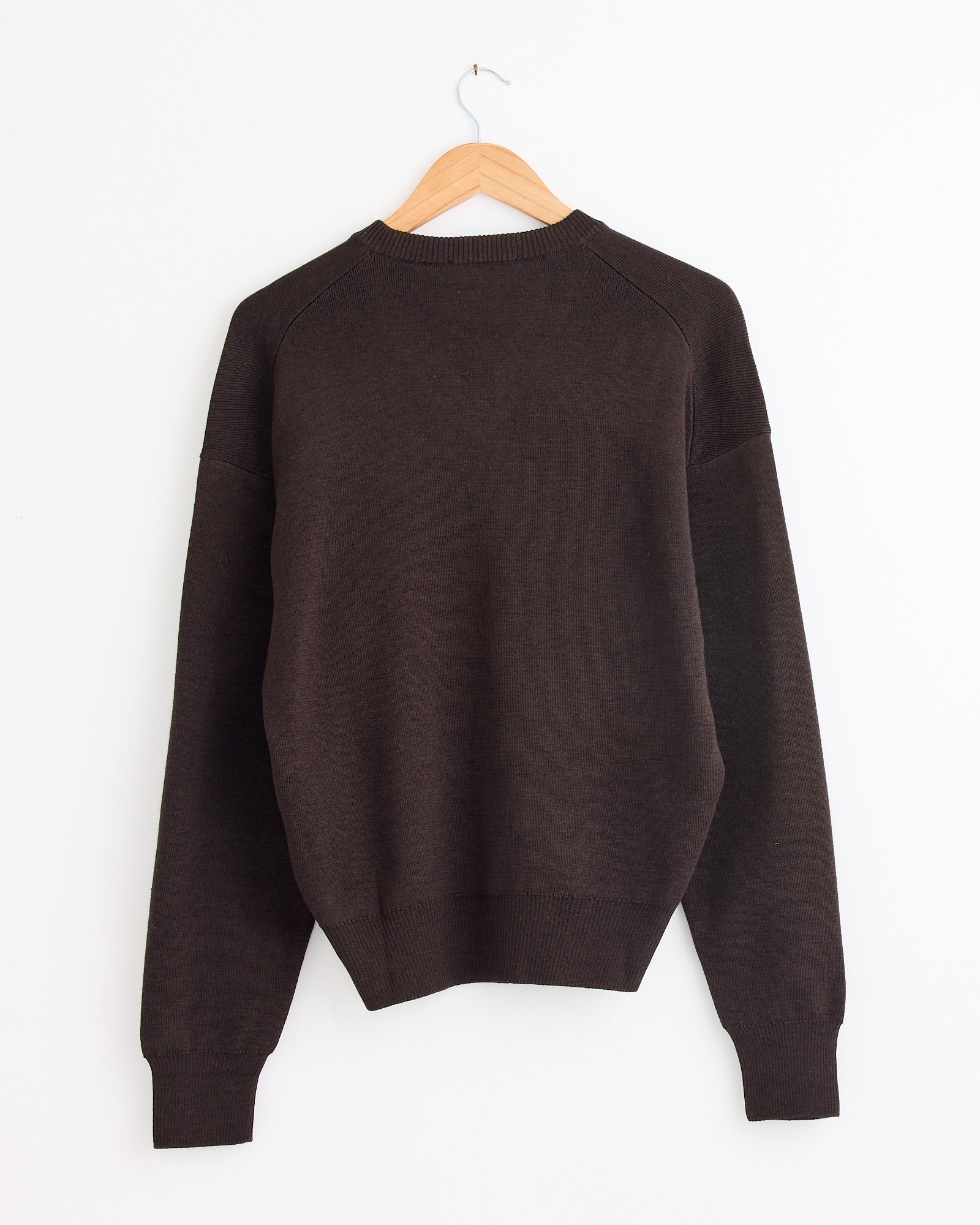 The Lemaire V Neck Sweater in Dark Cacao, featuring long sleeves, hangs on a wooden hanger against a plain white wall. For sizing or fit assistance, please use our online chat.
