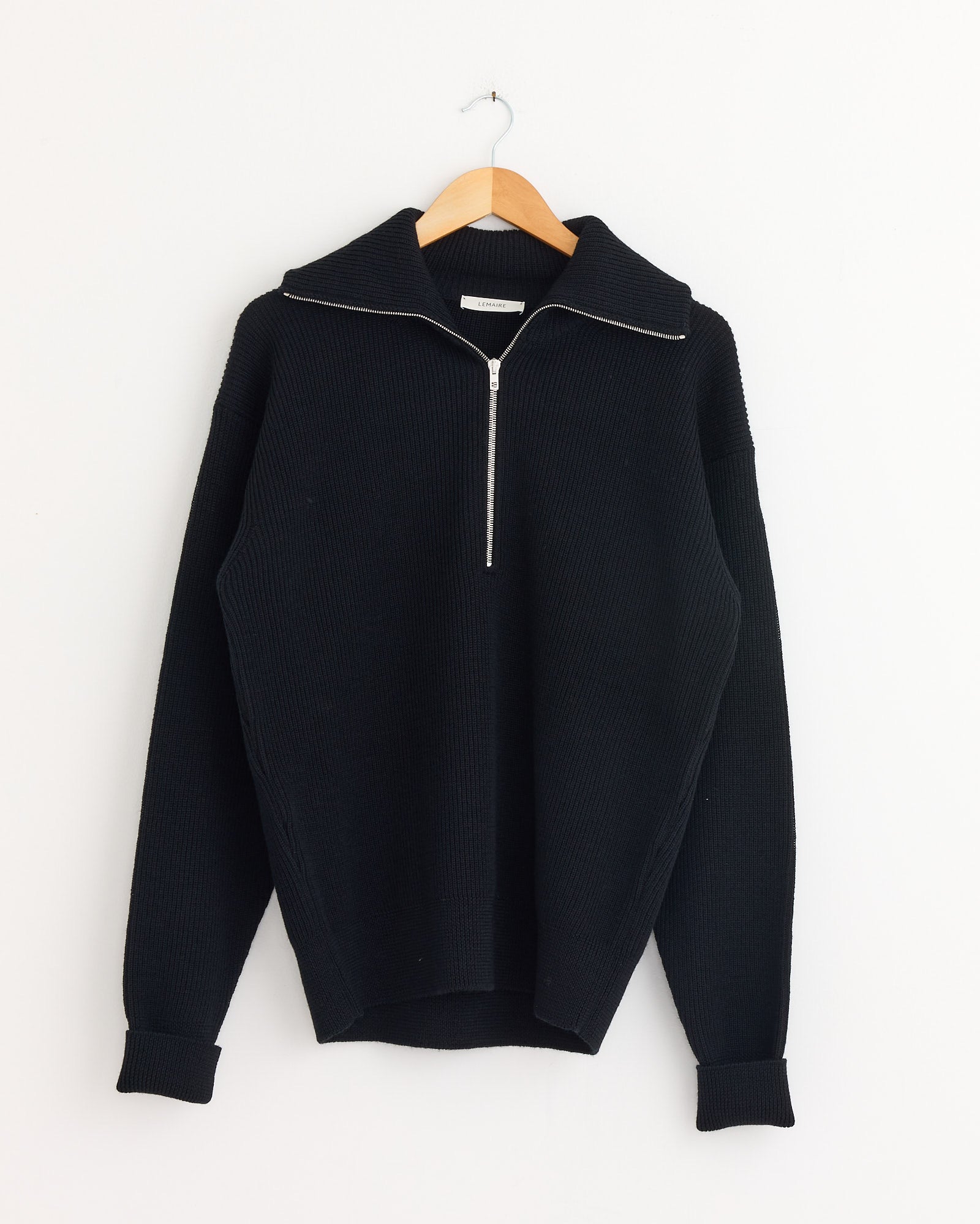 The Lemaire Half Zip Sweater in Black, crafted from ribbed virgin wool with long sleeves and a half-zip collar, hangs on a wooden hanger against a plain white wall.