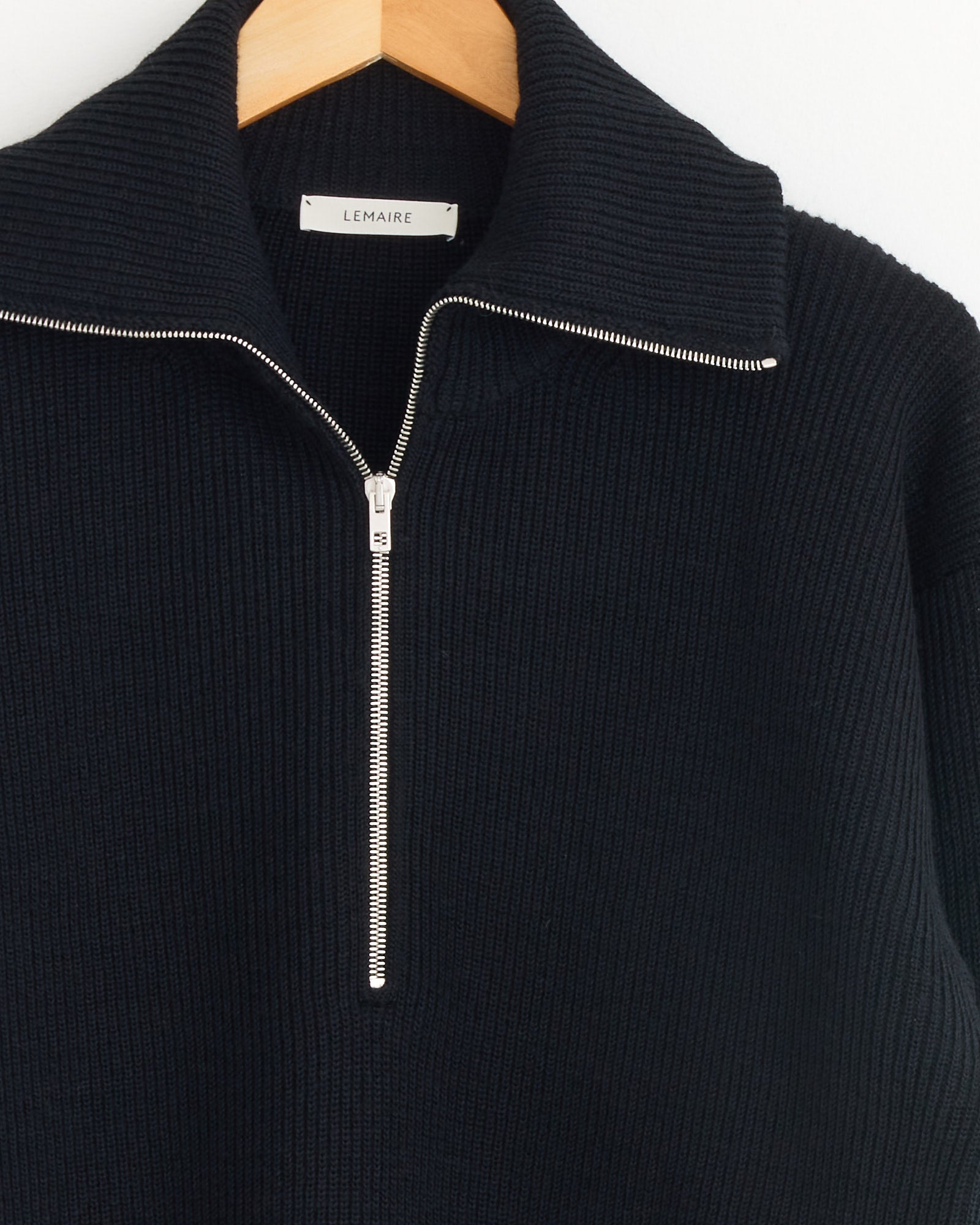 A close-up of the Lemaire Half Zip Sweater in Black, featuring ribbed virgin wool, a silver half-zipper, and wide collar, hanging on a wooden hanger with a label that reads LEMAIRE.
