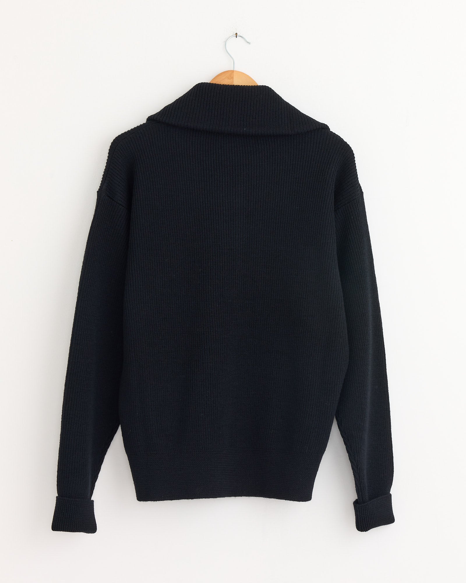 The Lemaire Half Zip Sweater in Black, crafted from ribbed virgin wool, hangs on a wooden hanger against a plain white wall, displayed from the back with slightly folded cuffs.