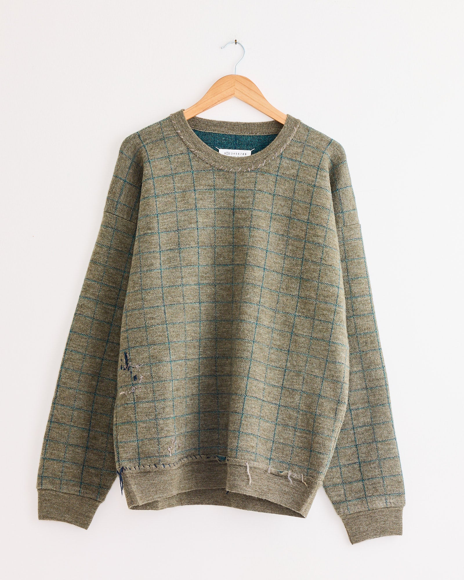 The Maison Margiela Crewneck in Green hangs on a wooden hanger against a white wall, featuring visible distressed details and minor holes near the hem.
