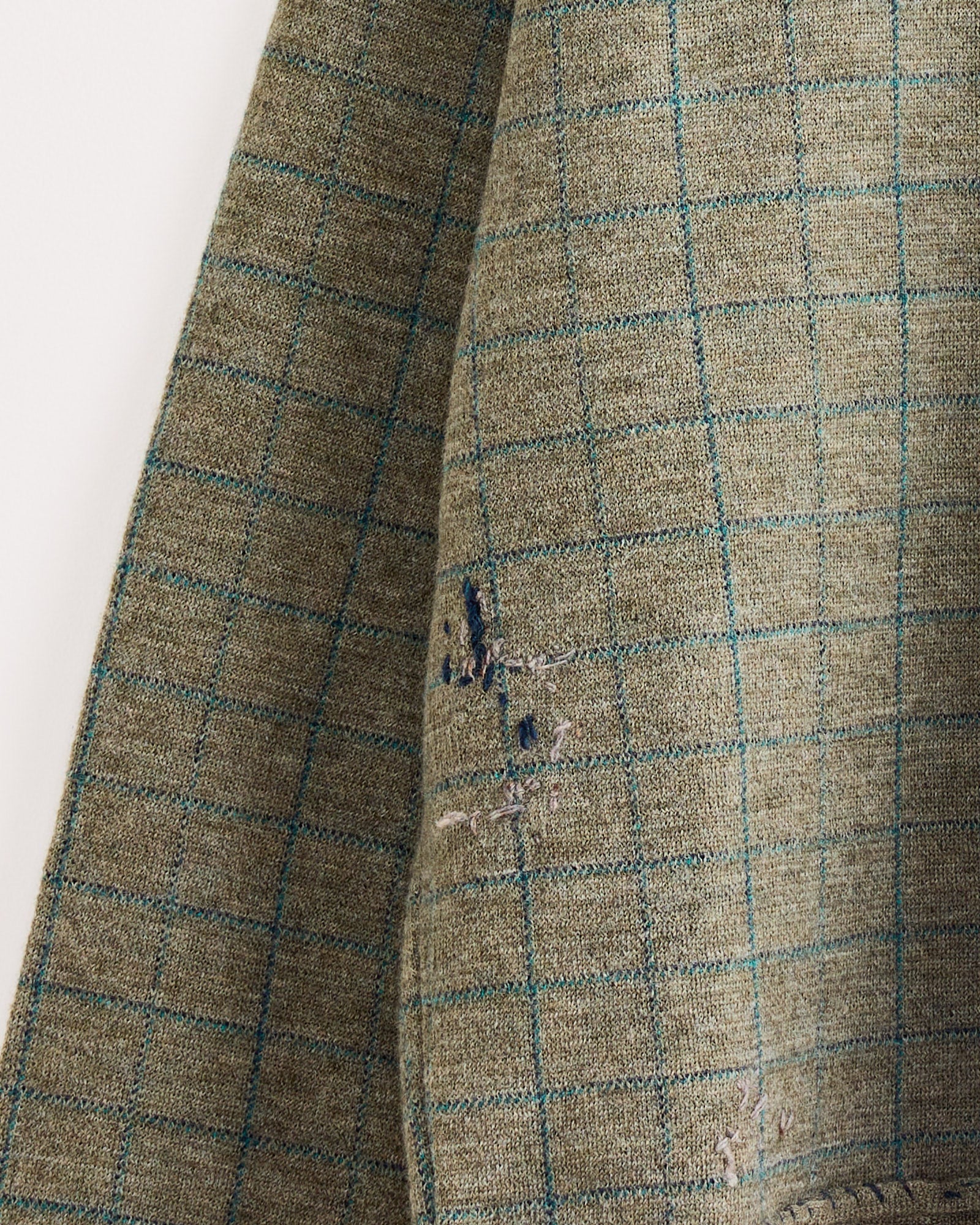 A close-up of the Maison Margiela Crewneck in Green, showing its blue checkered pattern and visible mending stitches on a worn area.