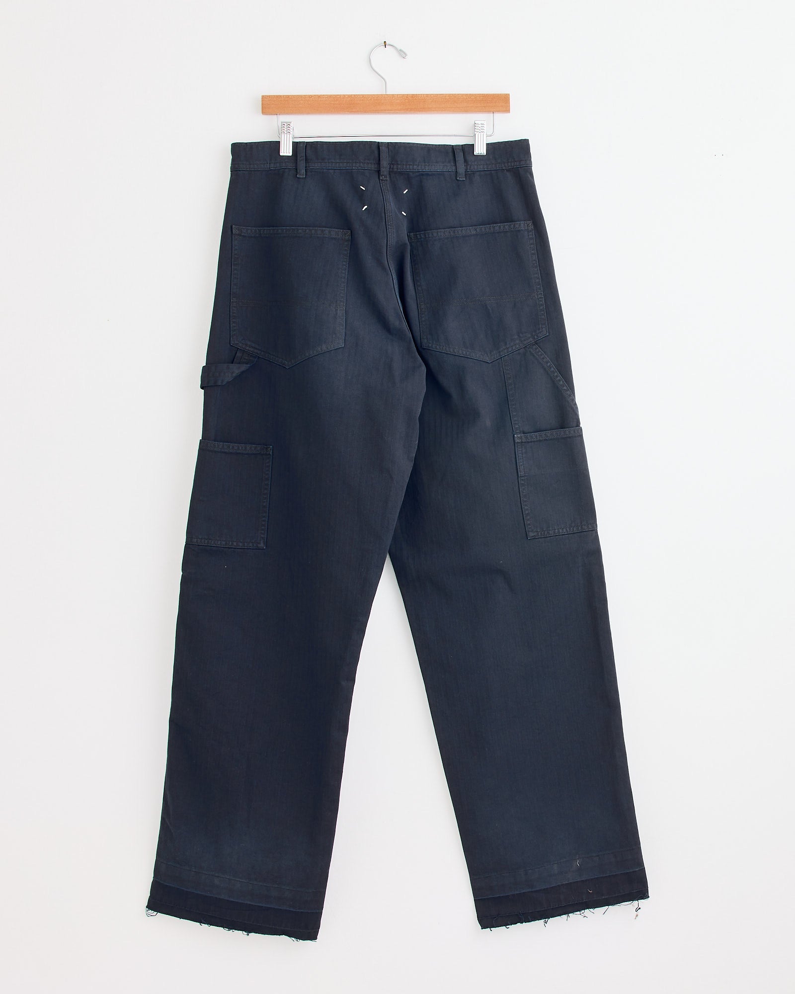 A pair of 5 Pockets Pant in Blue by Maison Margiela, featuring large back and side pockets, raw frayed hems, and a relaxed fit, hangs on a wooden hanger against a white wall.