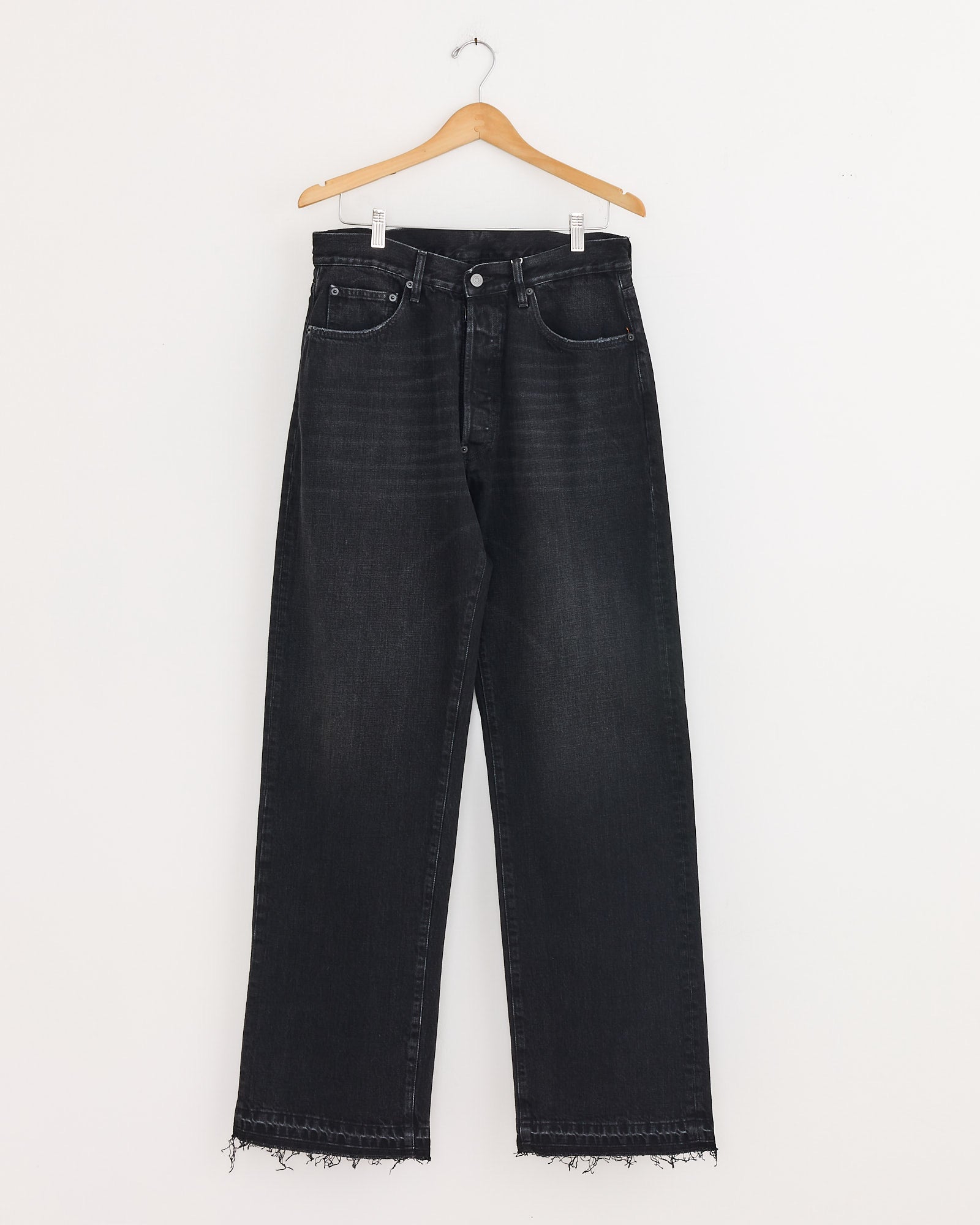 The Maison Margiela 5 Pocket Pant in Black, made from cotton denim with a high waist, straight relaxed leg, and raw hems, hangs on a wooden hanger against a plain white wall.