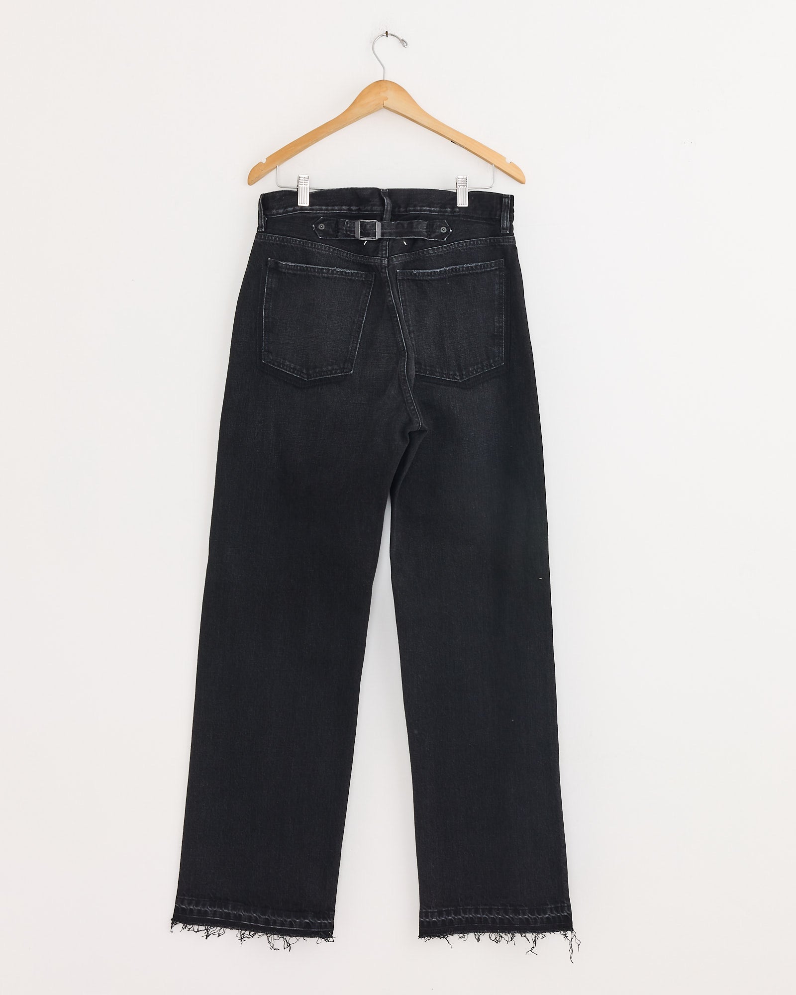 The Maison Margiela 5 Pocket Pant in Black, made from cotton denim, features a straight leg, high waist with belt loops, raw frayed hems, and two back pockets. Displayed on a wooden hanger against a white wall.
